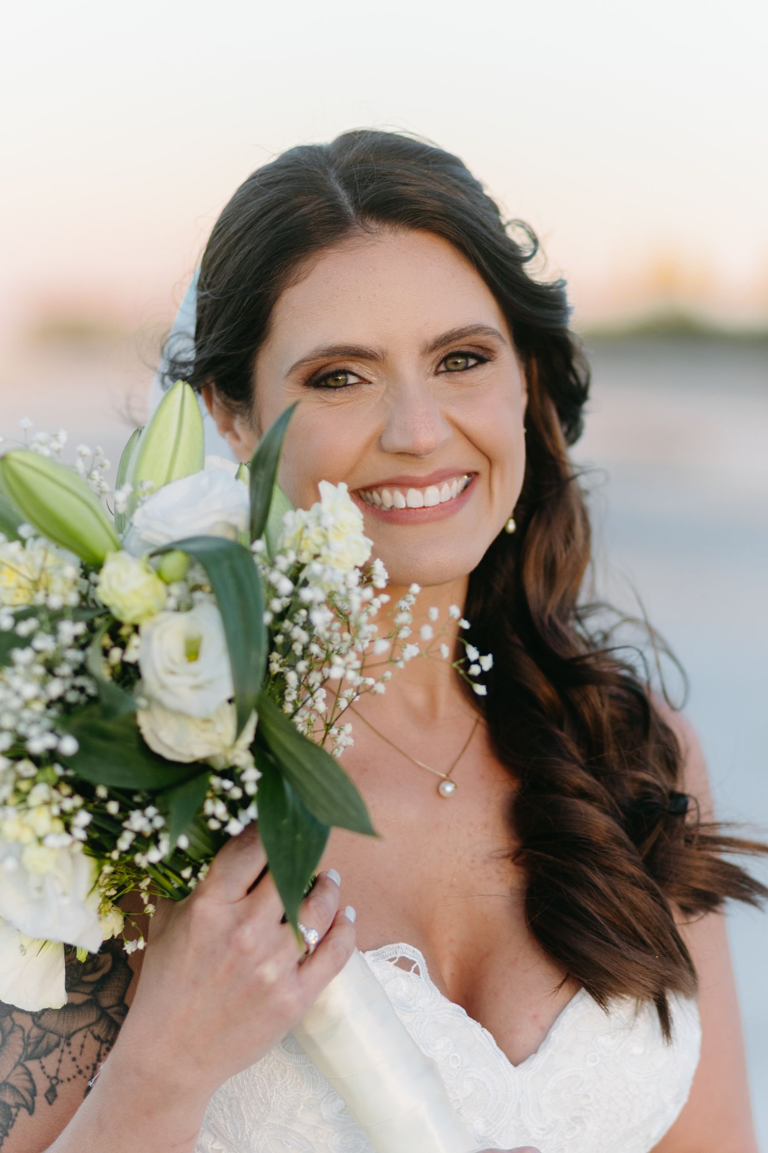 Bride in white lace dress smiles while holding white lily bouquet with baby's breath flowers against beach backdrop.