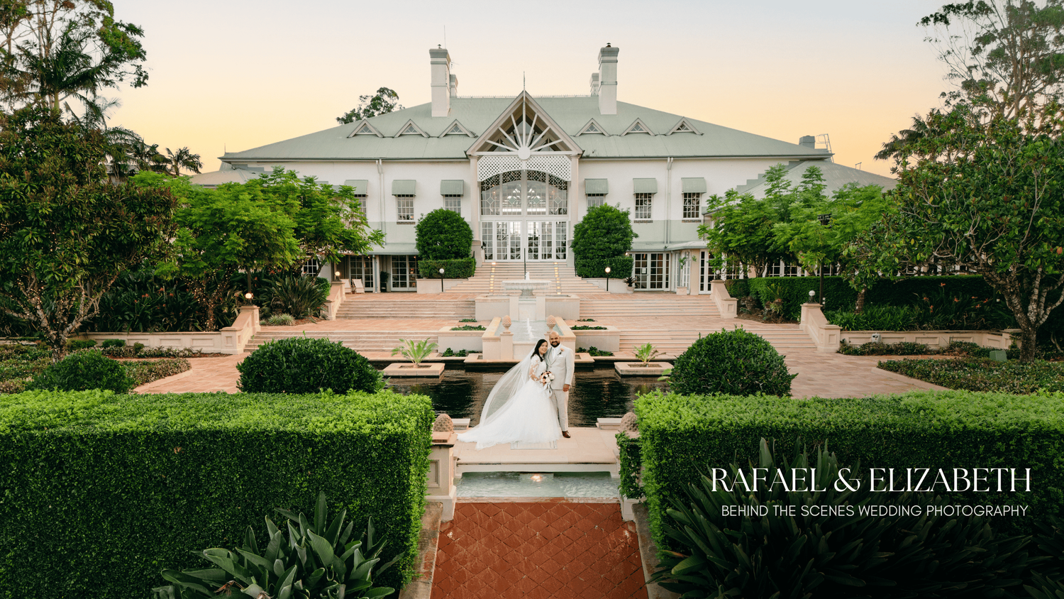 Elegant white mansion with manicured gardens and brick pathway leading to grand entrance at sunset.