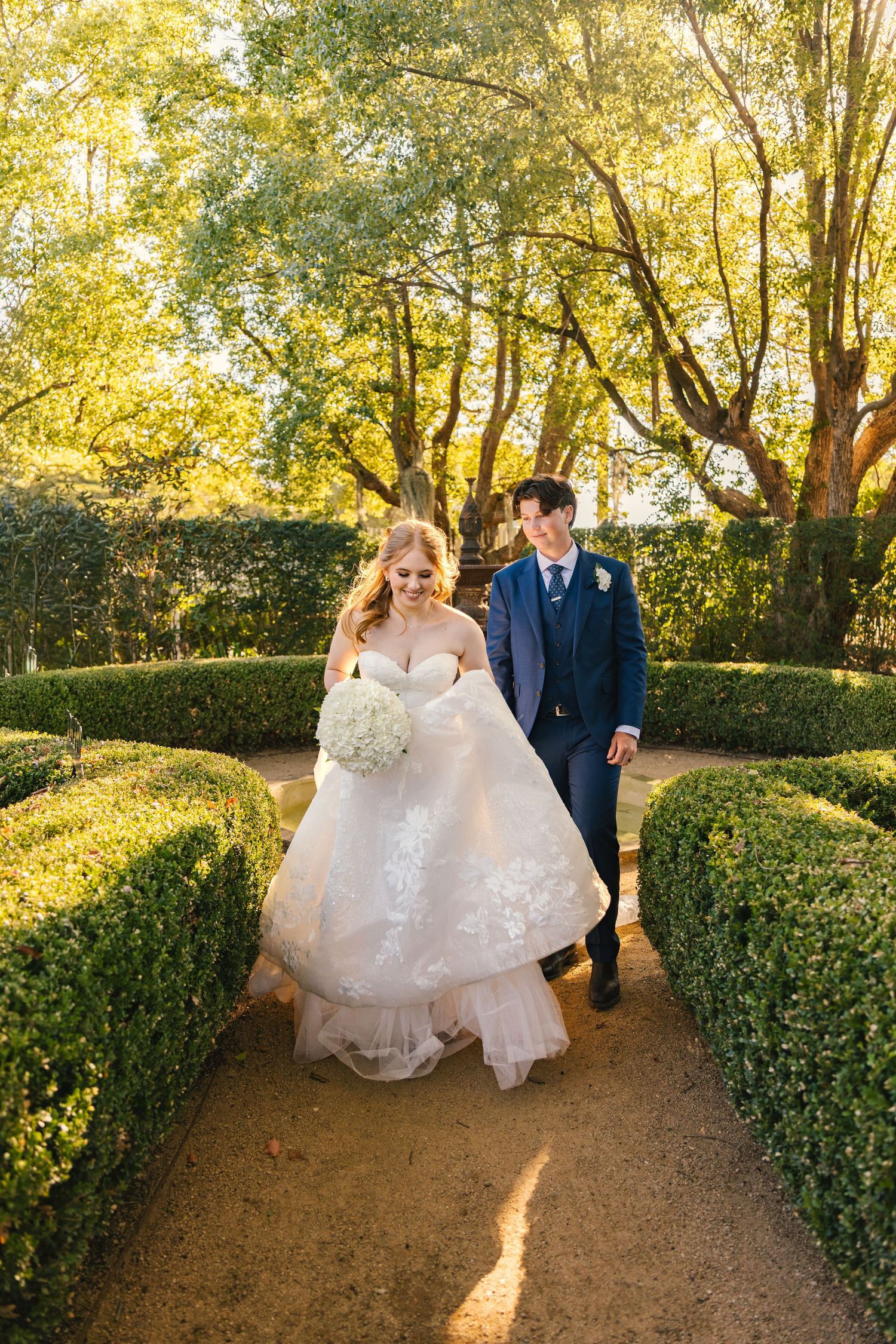 Bride in lace ballgown and groom in navy suit walk through manicured garden hedges on their wedding day.