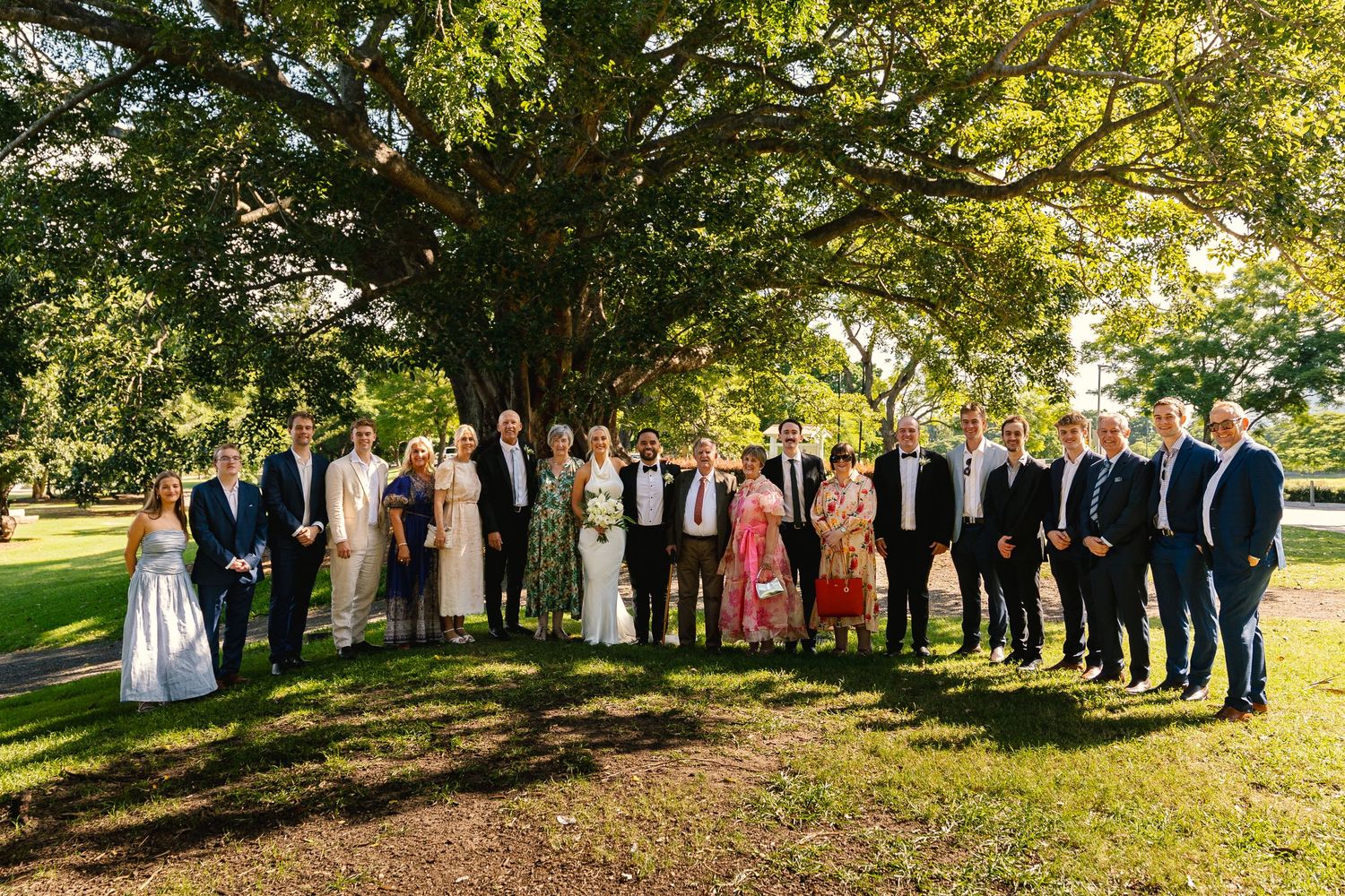 Wedding party members stand in a line outdoors under sunlit trees during a fall celebration.
