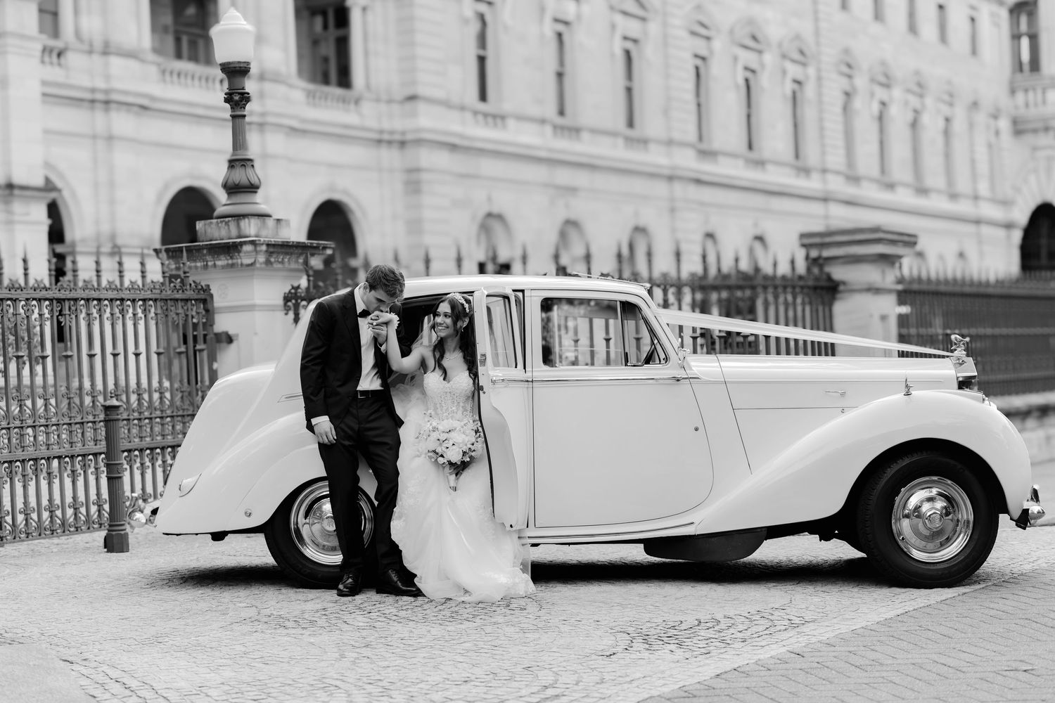 Bride and groom pose beside a vintage white Rolls-Royce decorated with ribbons outside a grand building.