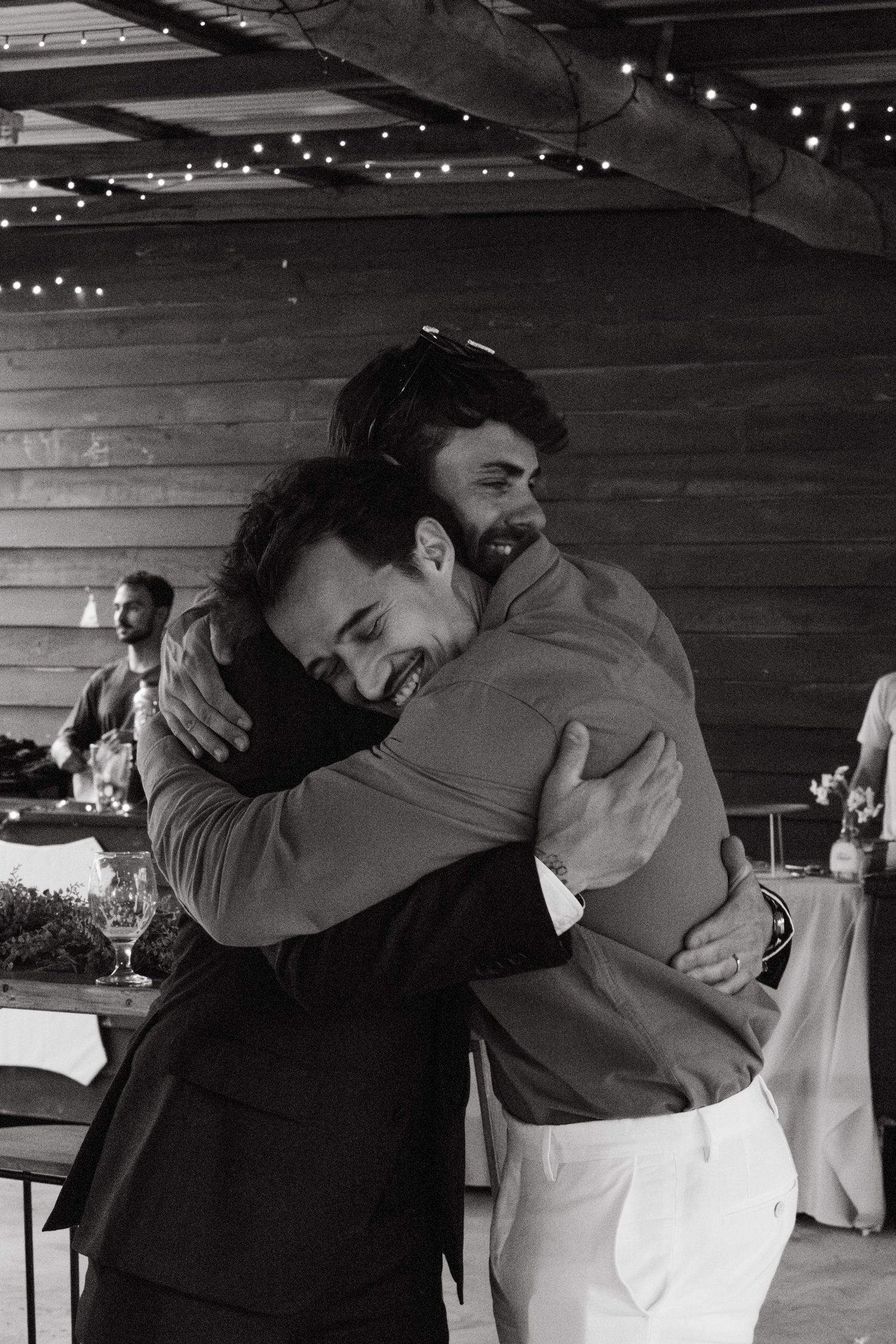 Two people embrace warmly in a rustic indoor venue decorated with string lights.