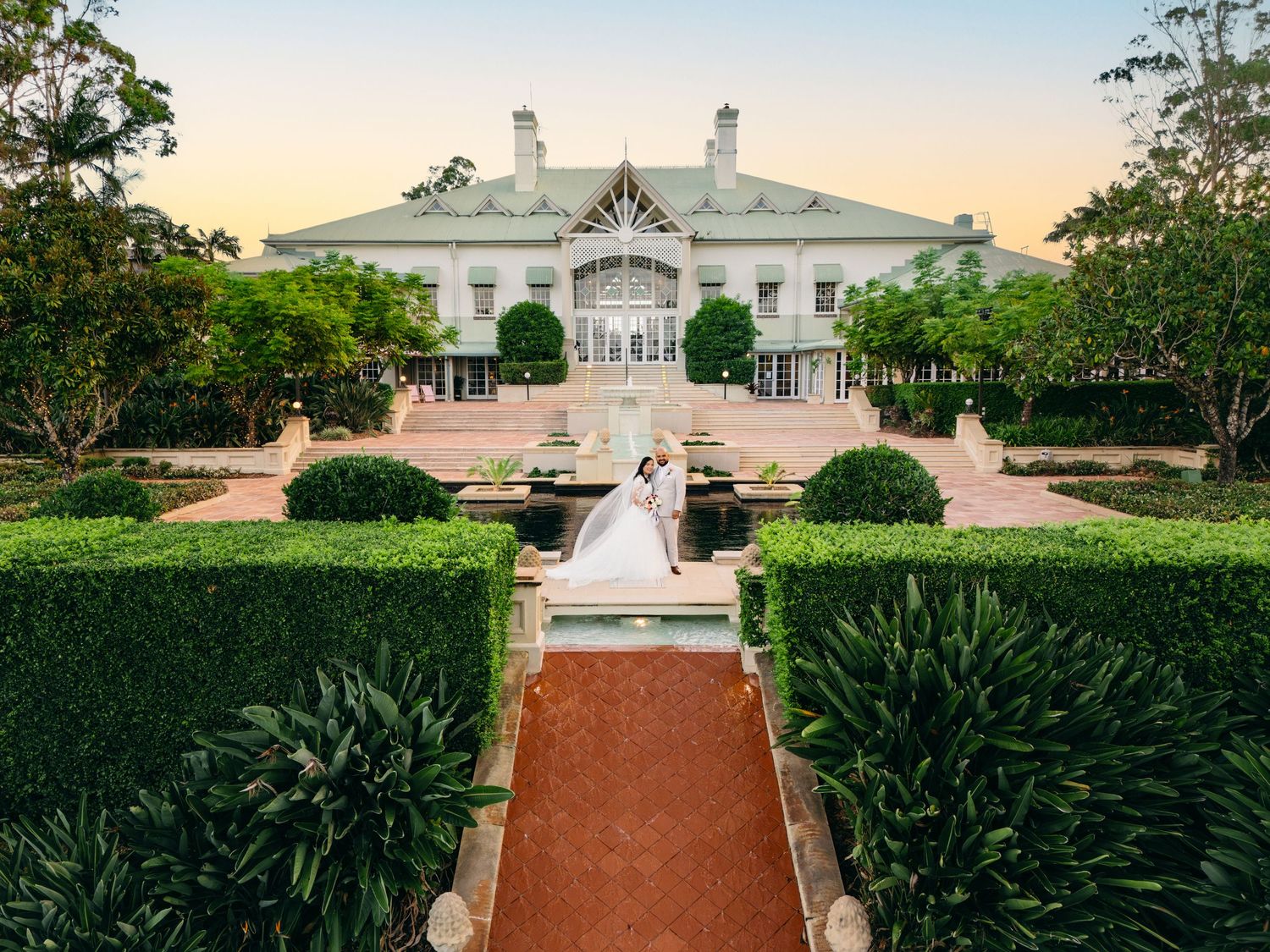 A panoramic view of a grand white mansion with manicured gardens and red brick pathways leading to its entrance.