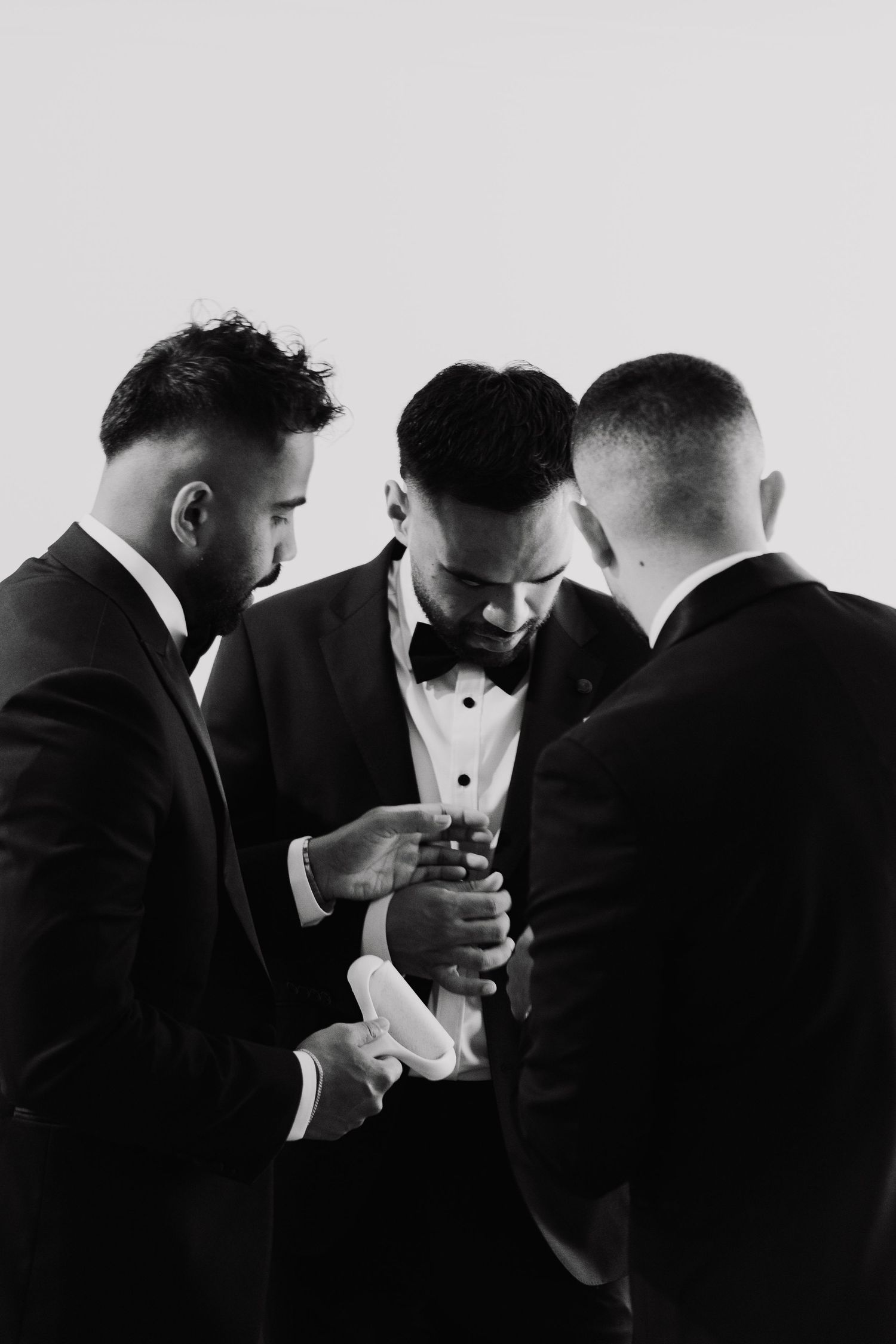Groomsmen in black tuxedos share a lighthearted moment while lighting cigars in dramatic black and white photography.