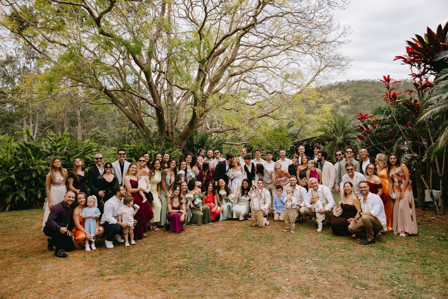 Large group of wedding guests pose together outdoors under trees for panoramic photo during daytime celebration.