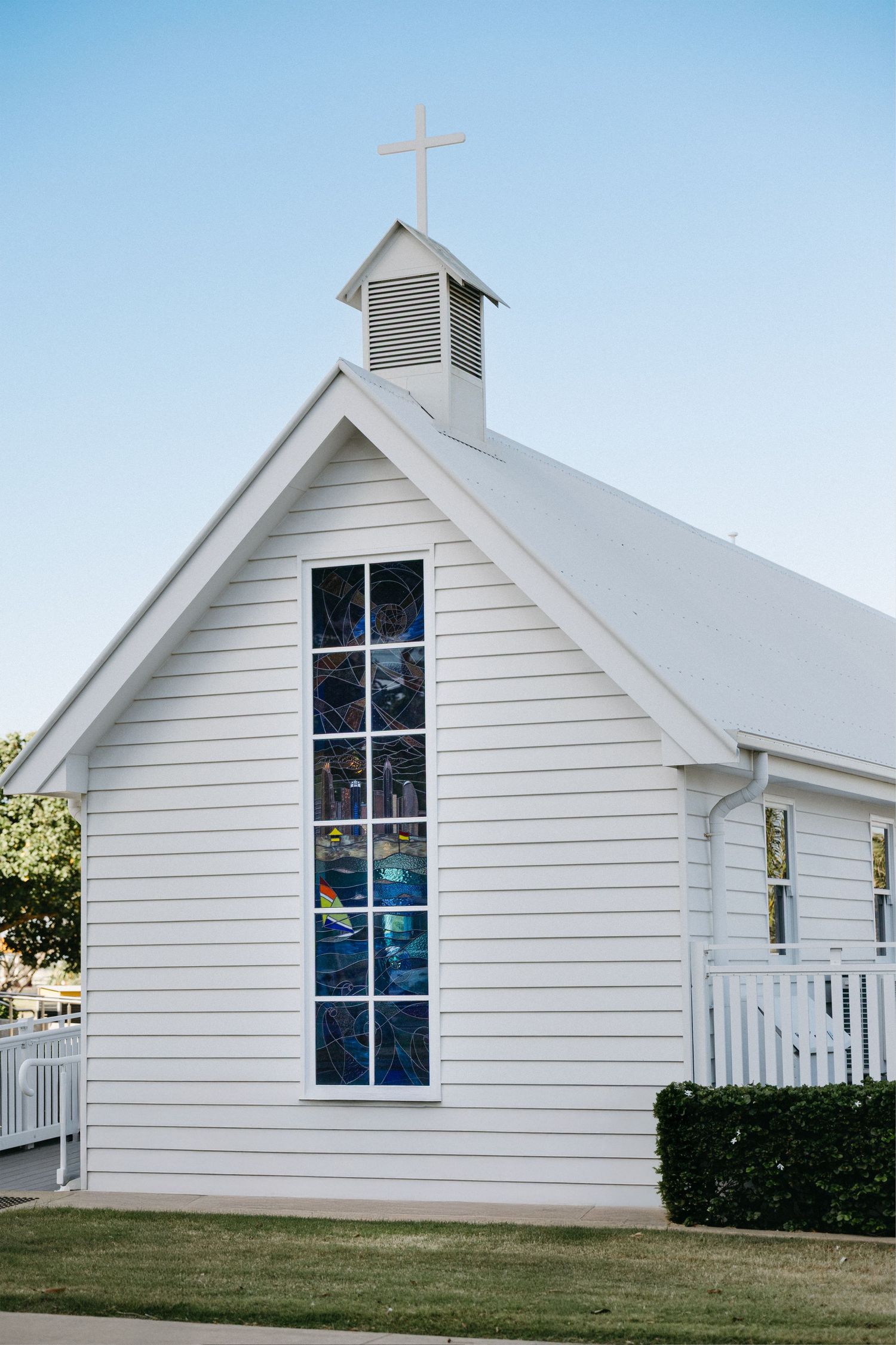 Small white chapel with tall cross-shaped stained glass windows and steeple photographed from different angles in daylight.