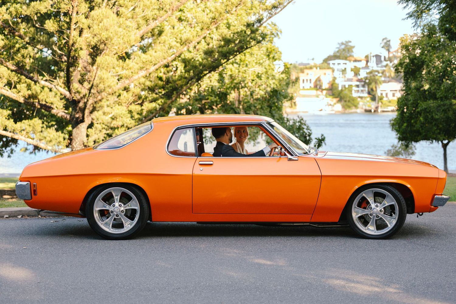 An orange vintage muscle car driving along a waterfront road on a sunny day.