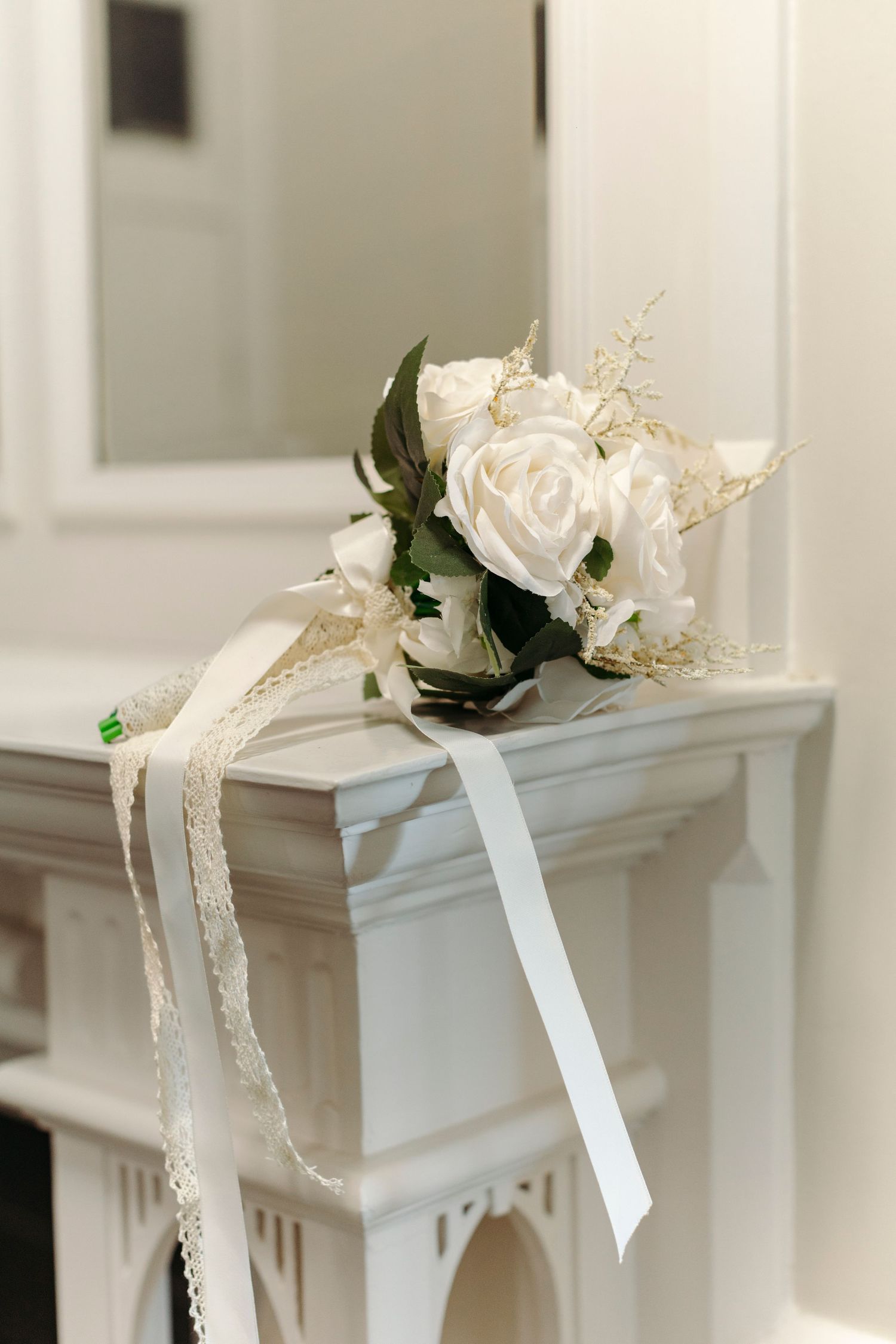White decorative floral arrangements with ribbons adorn a classic white fireplace mantel in an elegant interior setting.