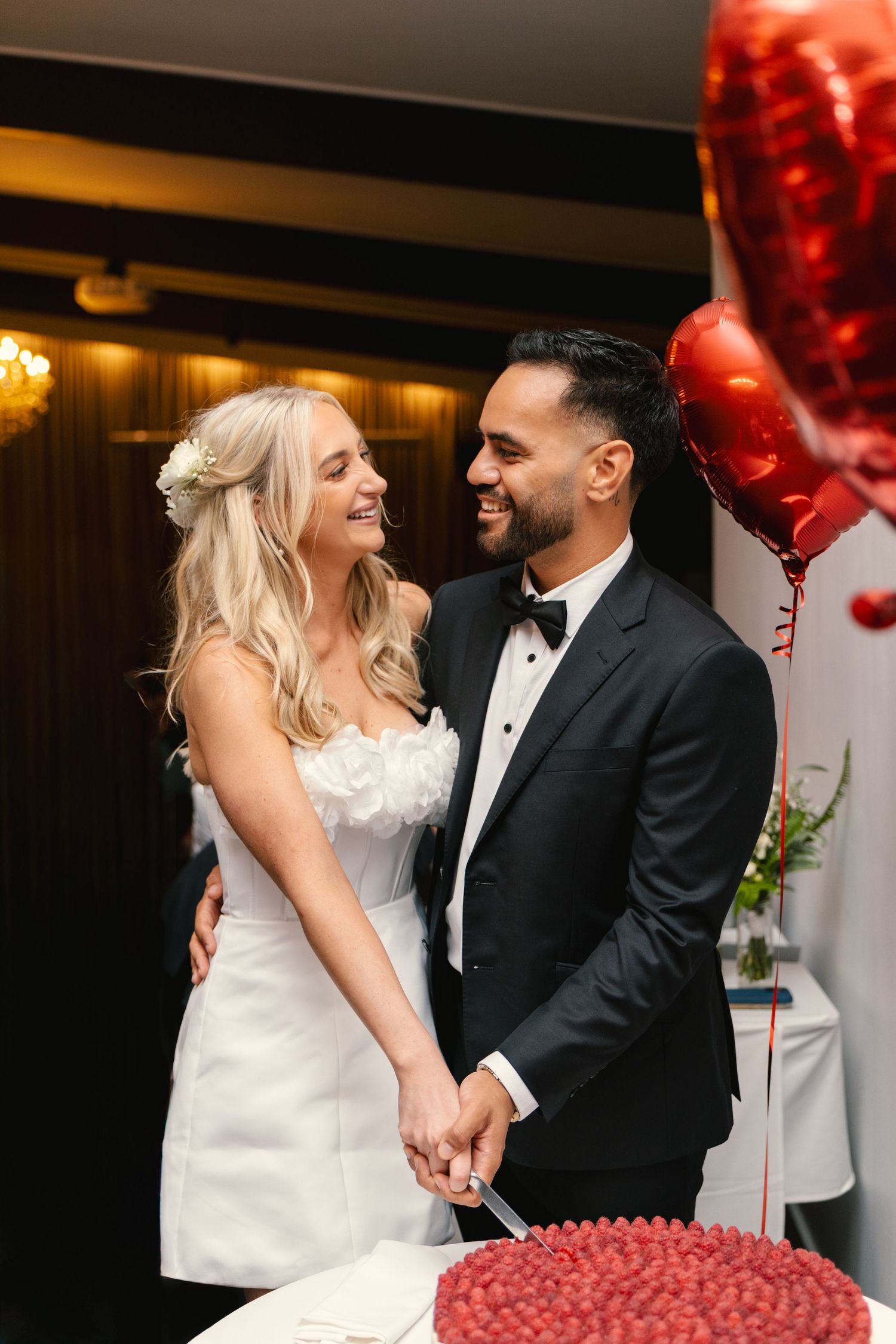 A couple shares an intimate moment at their wedding celebration with red heart balloons in the background.