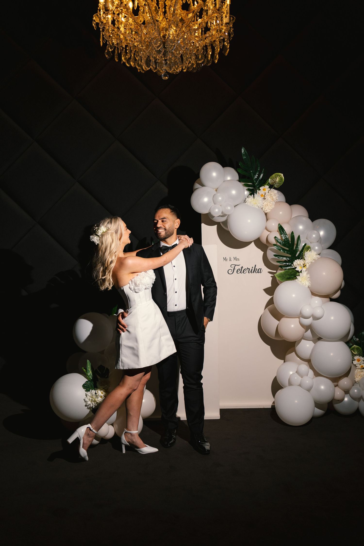 Elegant wedding couple poses playfully against a backdrop of white balloons and a crystal chandelier.