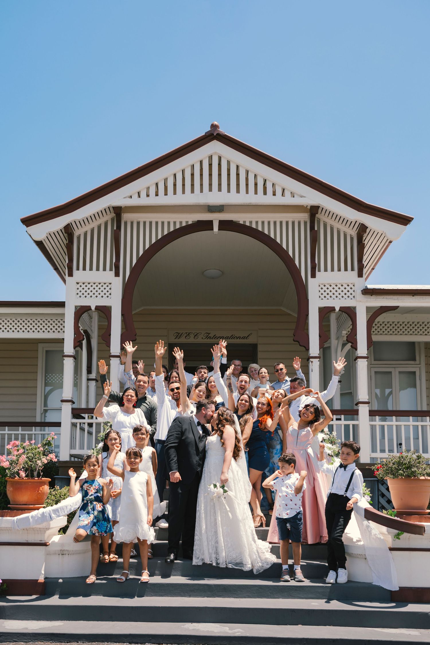 Wedding celebration group photo on the porch of a white colonial-style building with decorative trim.
