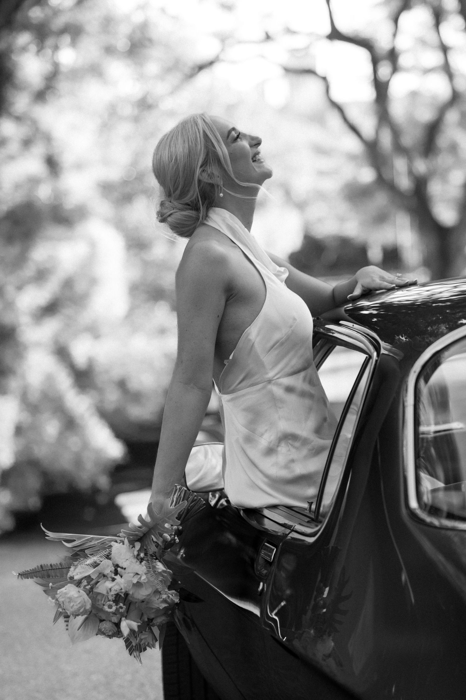 Bride in white dress leans against vintage black car while holding bouquet in black and white photography.