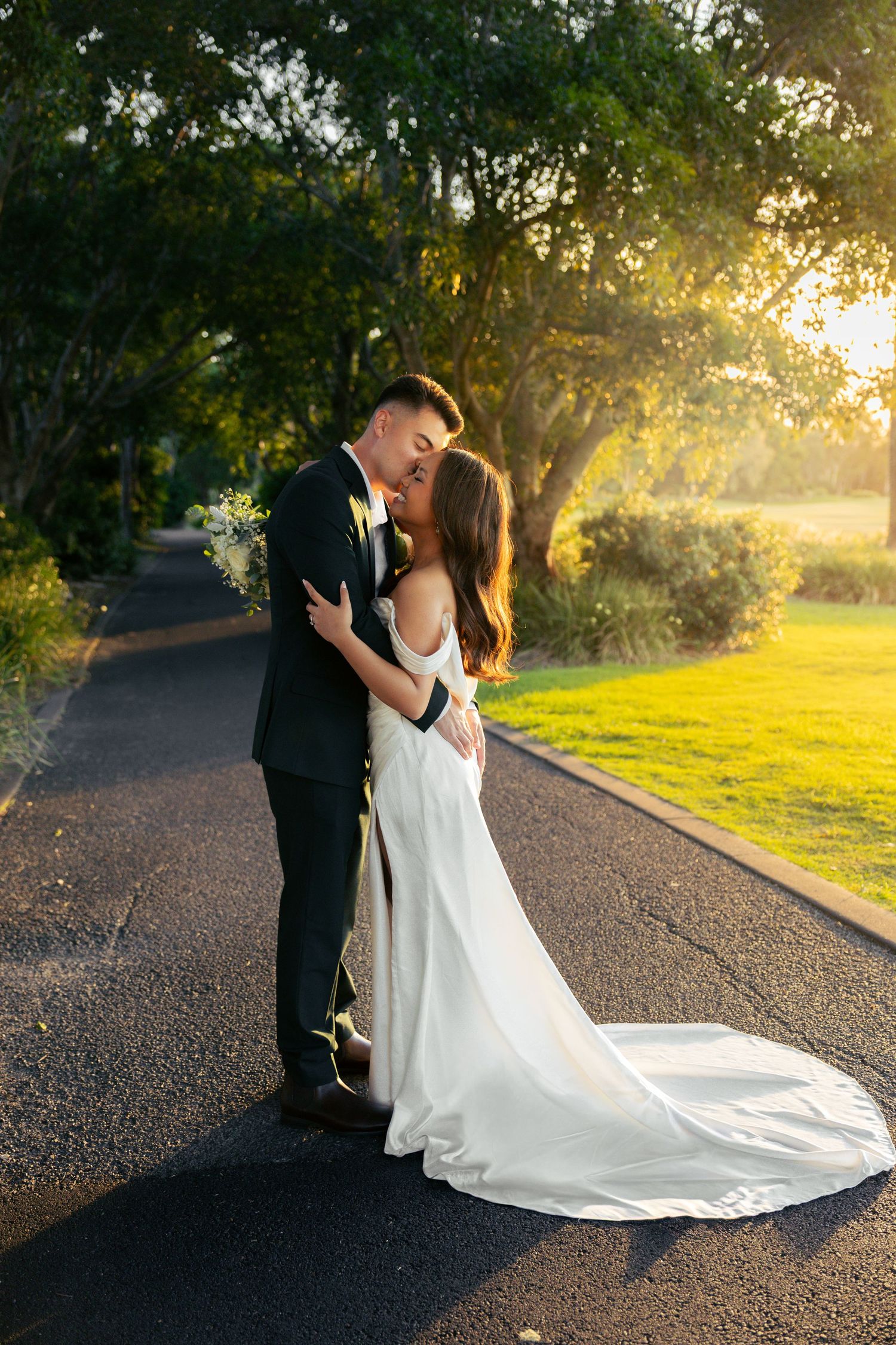 A romantic wedding couple embraces on a sunlit path during golden hour with a flowing white dress trailing behind them.