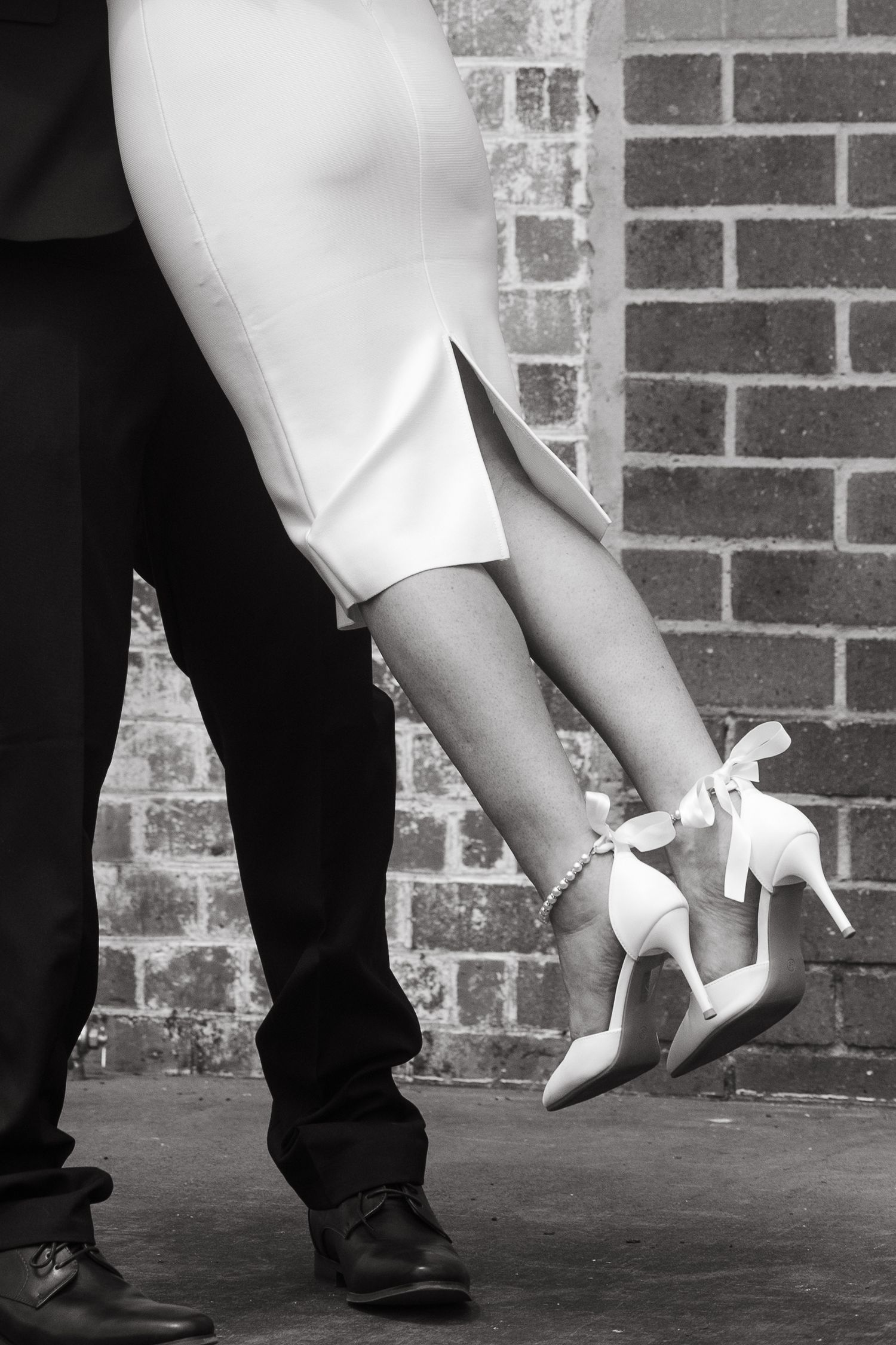 Black and white photo showing dancing feet with one person wearing white shoes and another in dark shoes against brick wall.