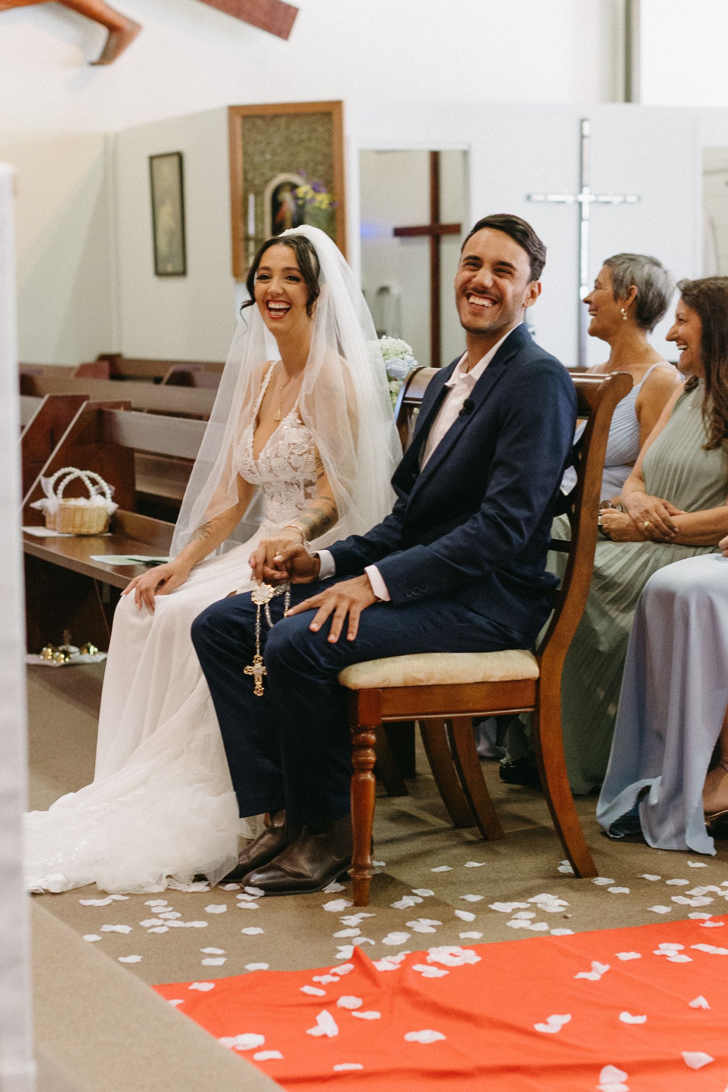 Wedding party sitting and laughing together on church pews with rose petals scattered on a red carpet.