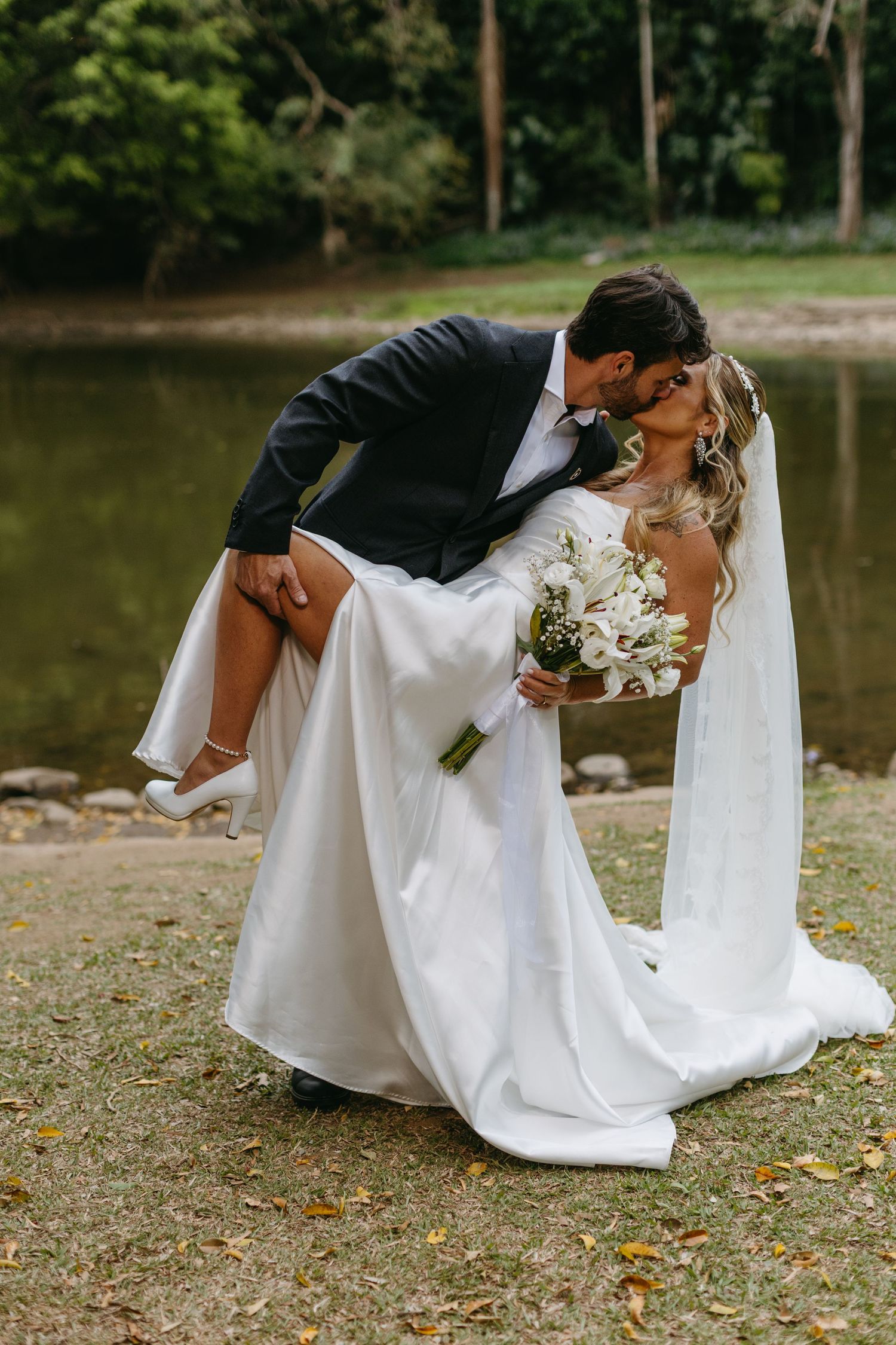 Romantic wedding couple poses by lakeside in dramatic dip pose.