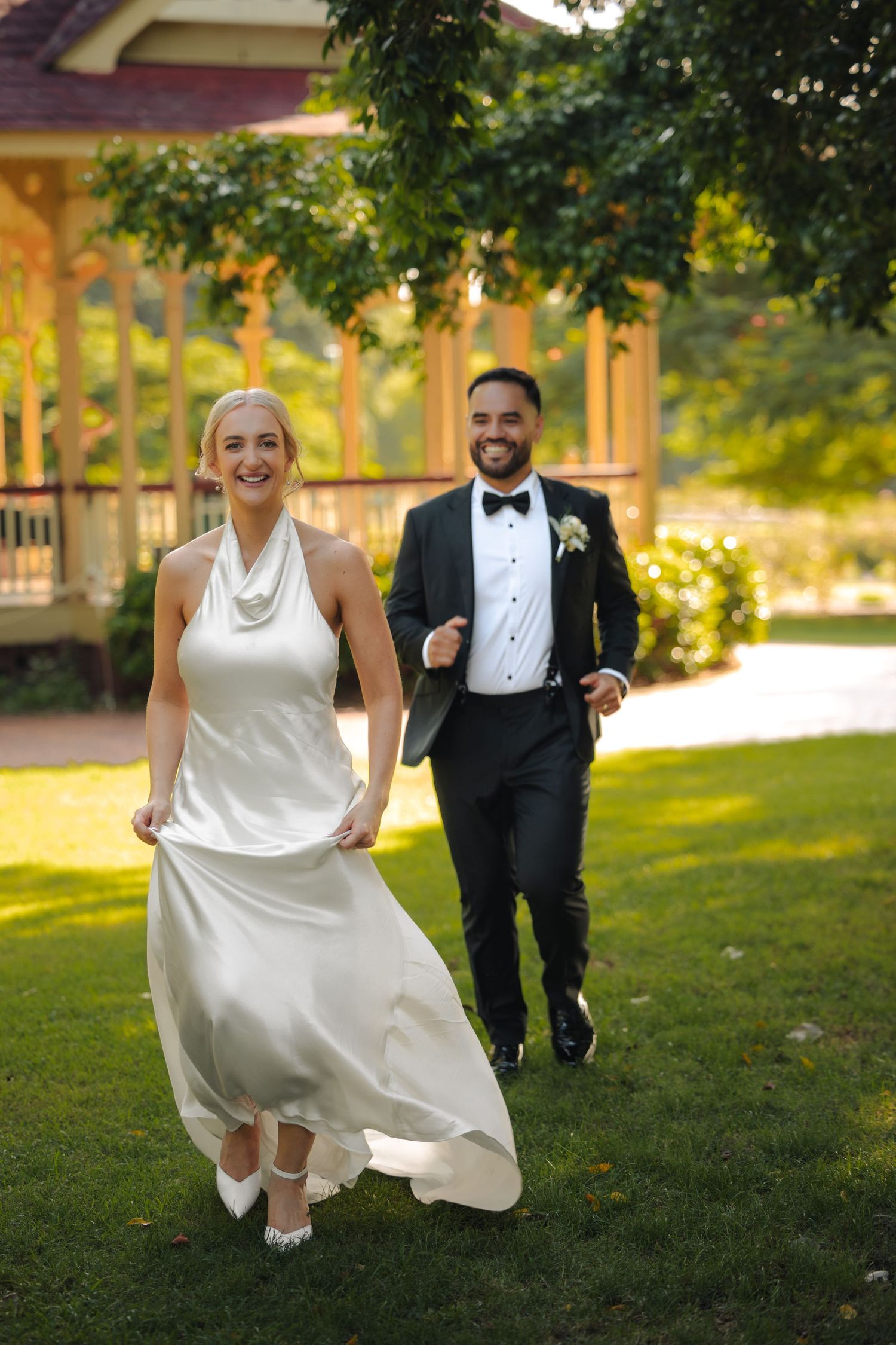 A joyful wedding couple playfully poses on a grassy lawn during golden hour with the bride lifting her white dress.
