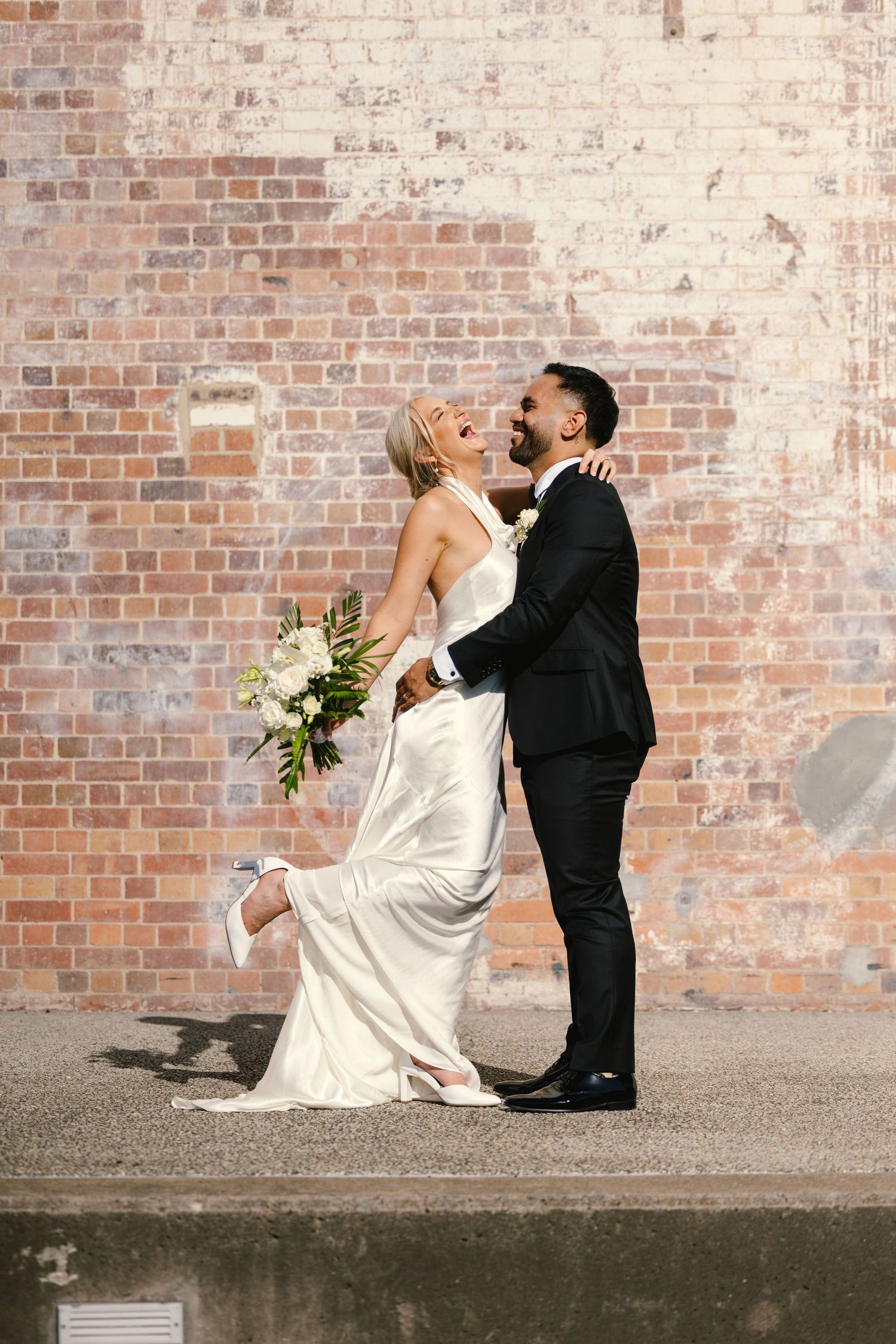 Newlyweds share romantic moment against rustic brick wall with bridal bouquet.