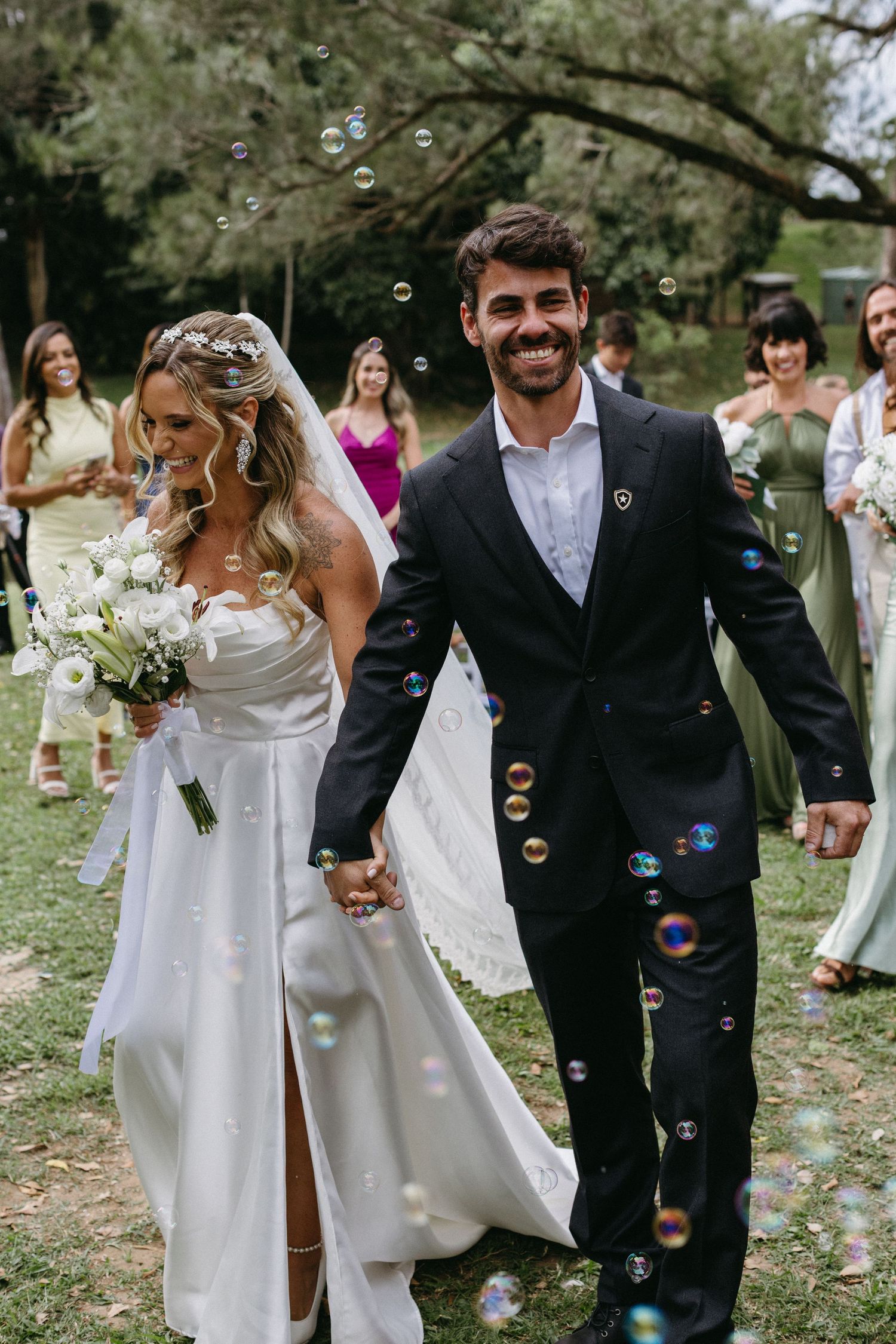 A joyful wedding couple walks through soap bubbles on a grassy lawn surrounded by guests.