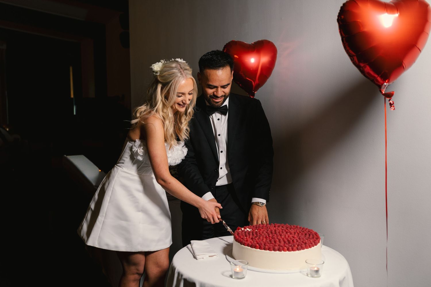 A wedding couple cuts their red and white wedding cake decorated with heart-shaped balloons at their reception.