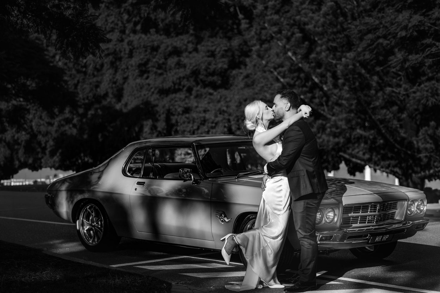 Black and white photo of a couple embracing and kissing beside a classic vintage car outdoors.