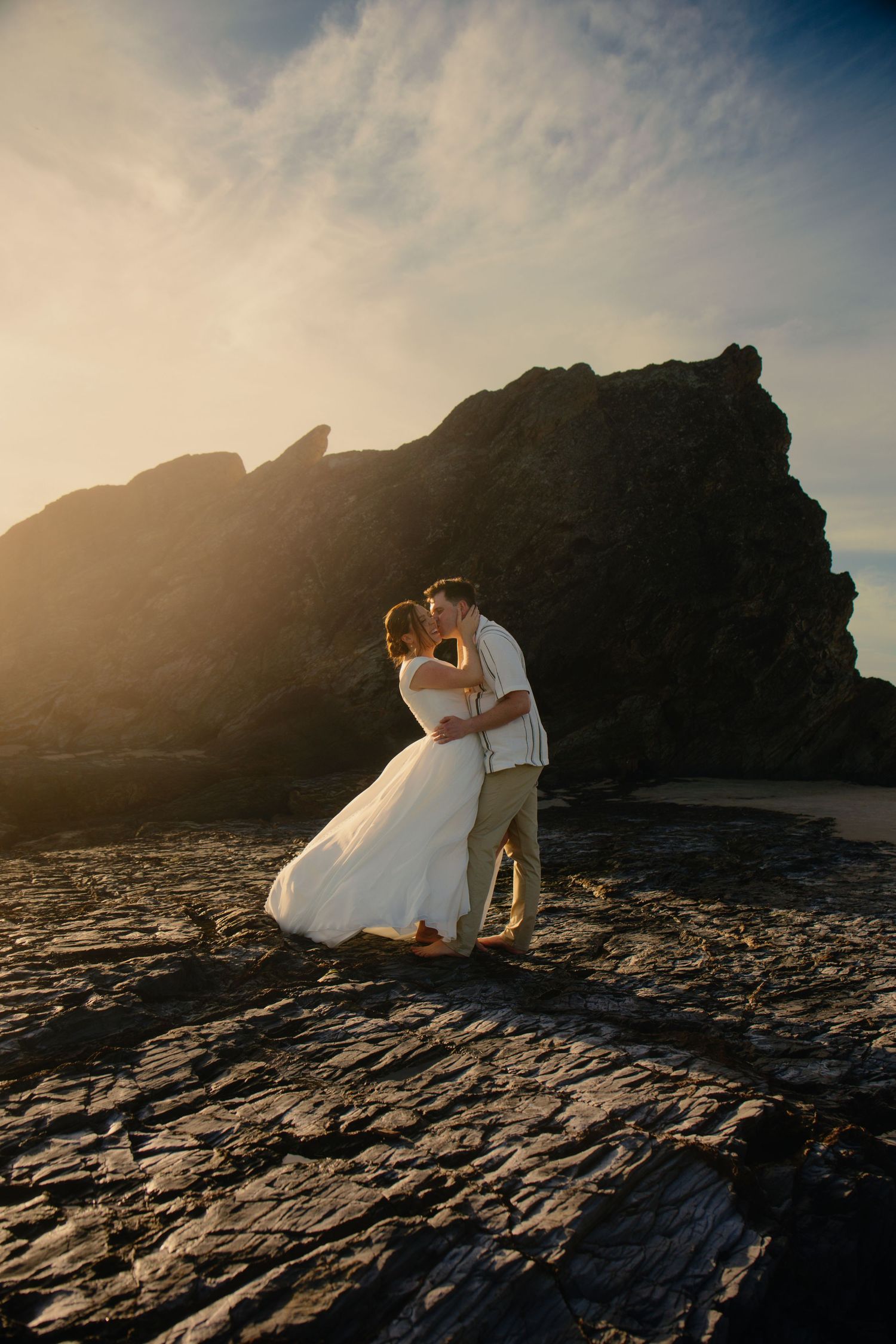 A couple sits on dramatic coastal rocks at sunset with mountains silhouetted in the background.