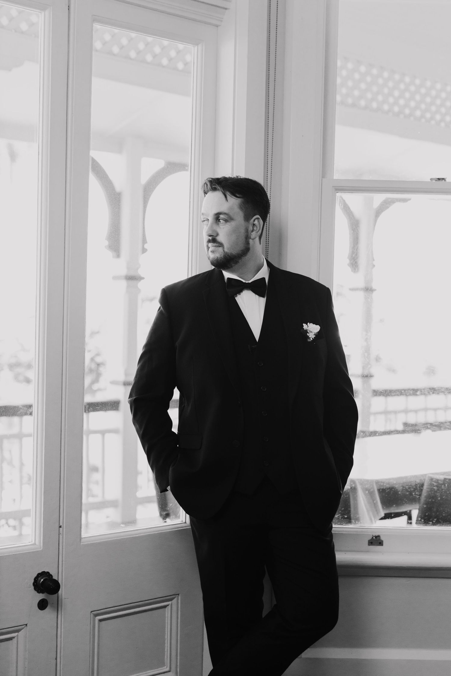 Formal portrait in black and white of someone in a tuxedo standing by ornate windows with decorative trim.