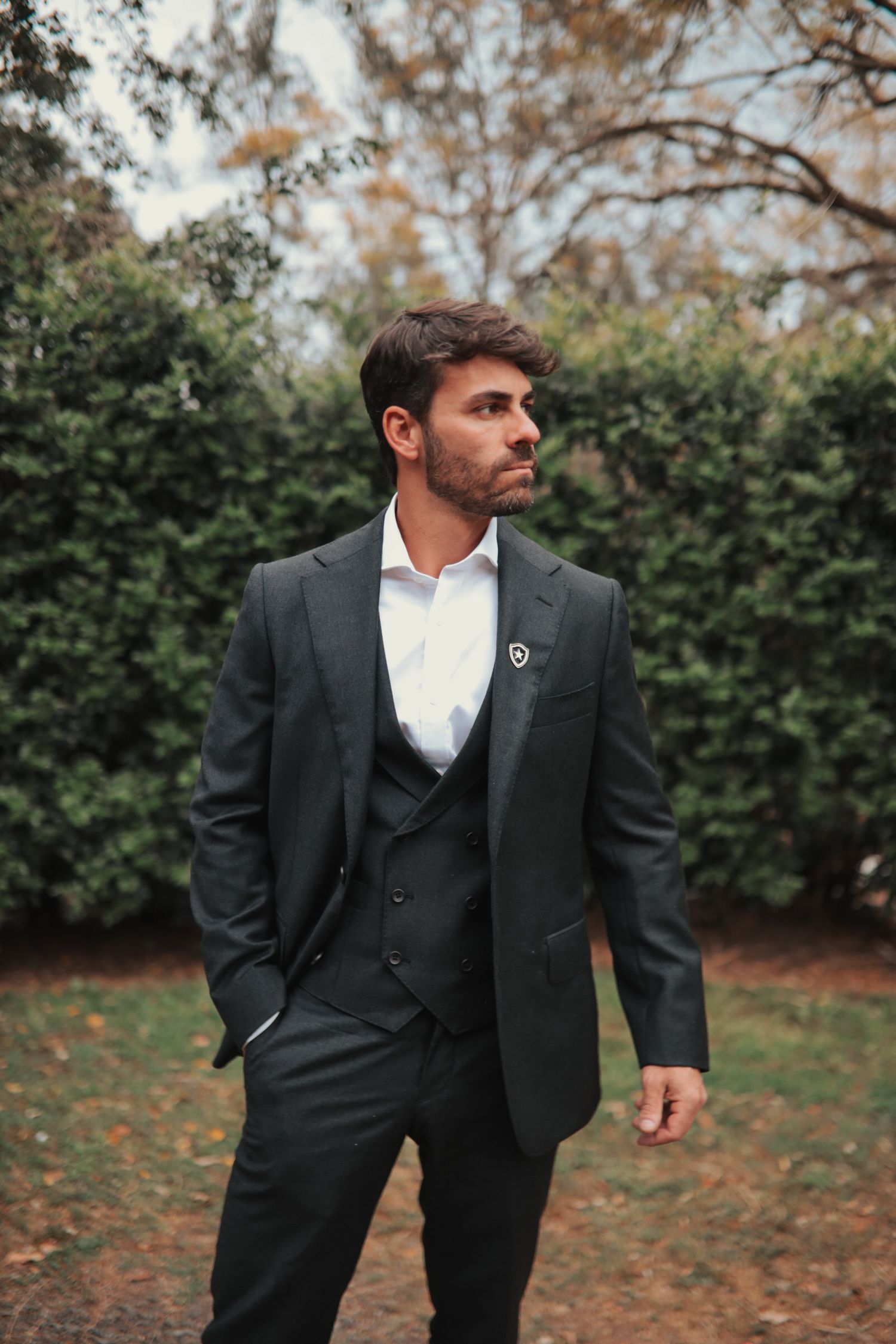 Groom in dark three-piece suit poses thoughtfully in an autumn garden setting.