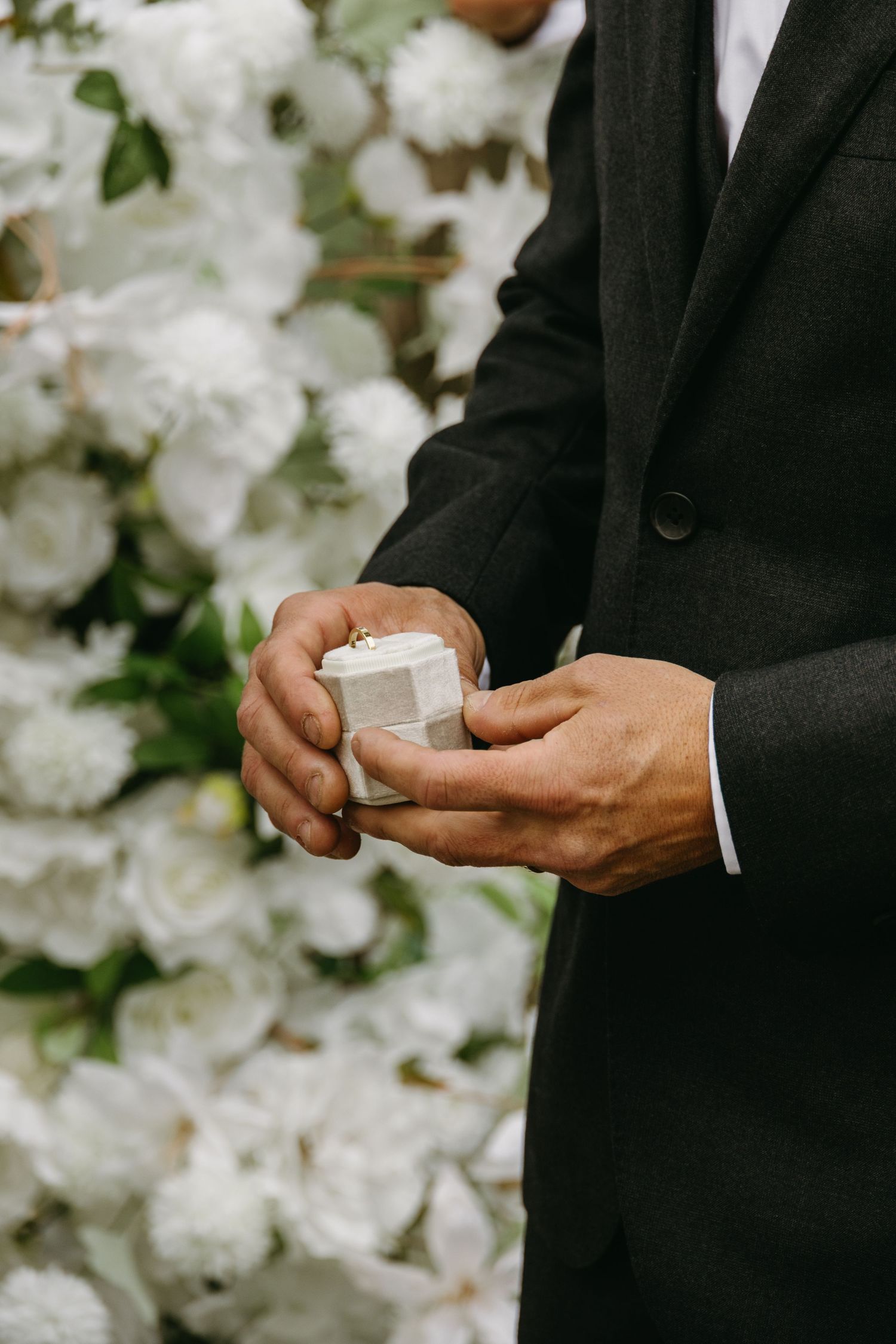 A person in a black suit holds a small white ring box against a backdrop of white floral decorations.