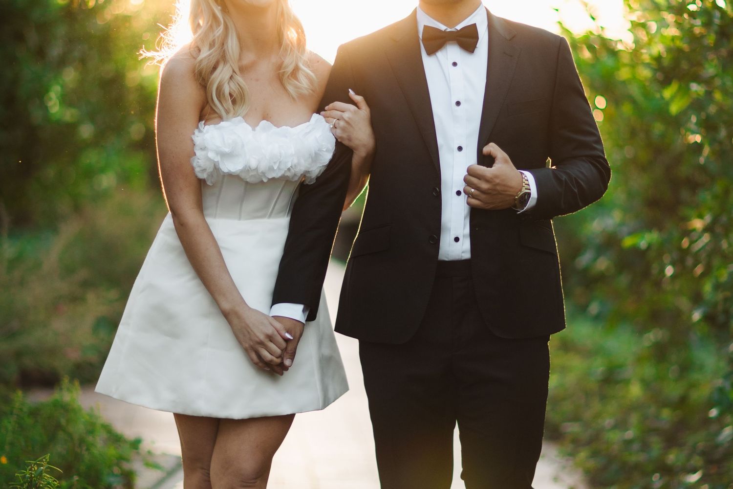 A couple in wedding attire walks together through a sunlit garden setting during golden hour.