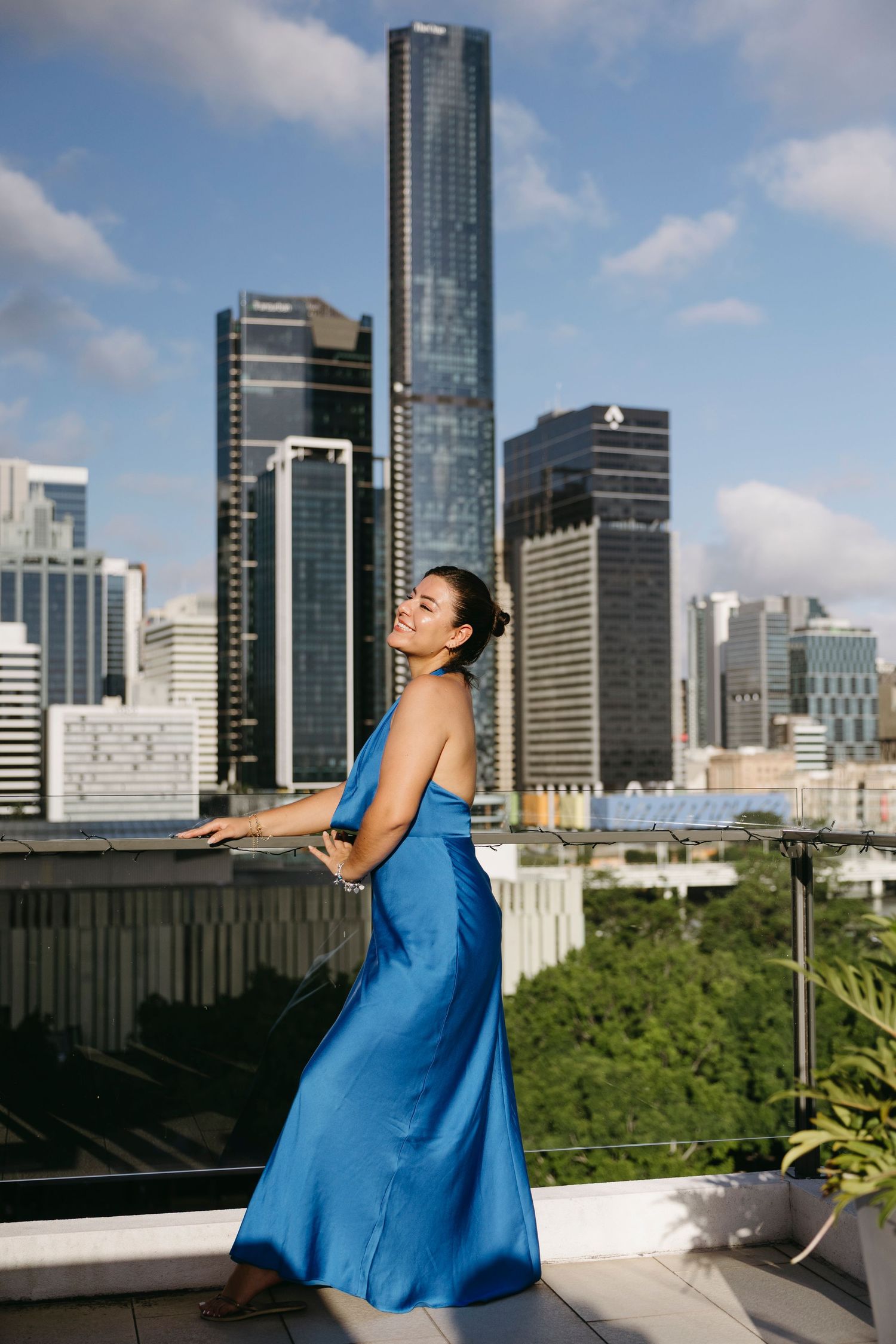 A person in a flowing blue dress poses on a rooftop terrace with Brisbane's modern skyline and high-rise buildings in the background.