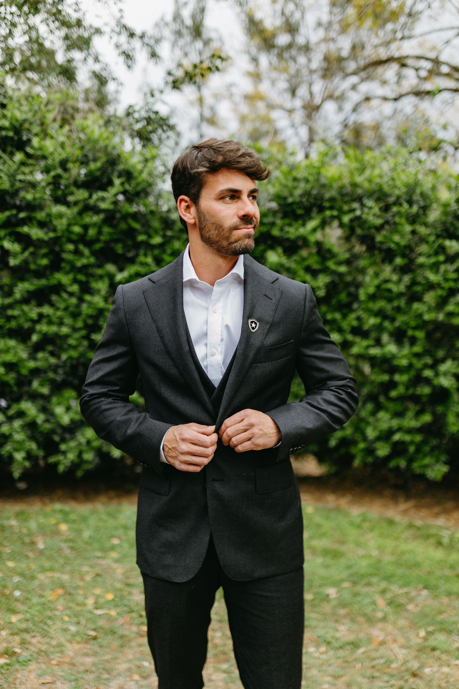 A man in a dark suit and white shirt adjusts his jacket while standing in front of a green hedge outdoors.