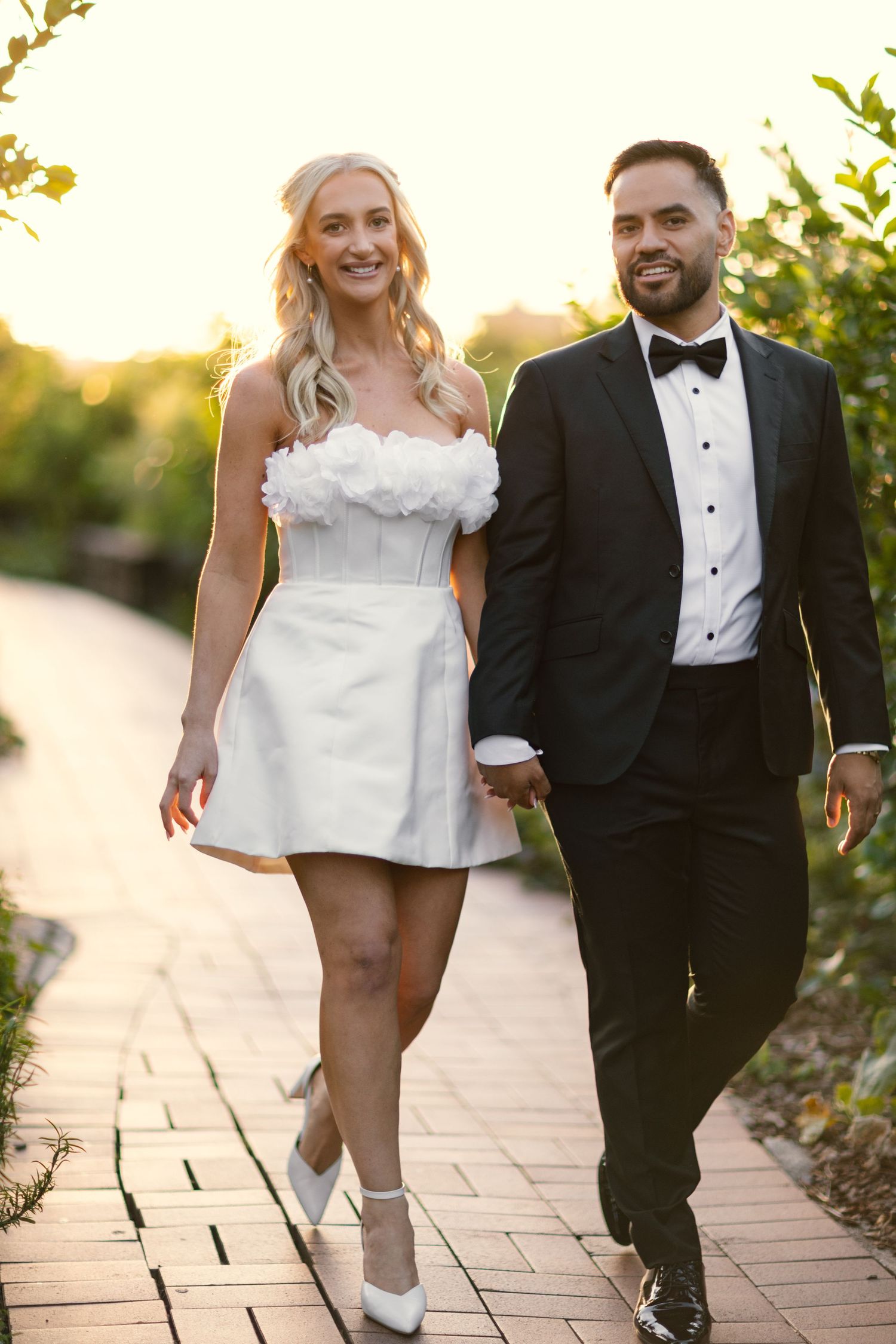 A couple in formal attire walks together on a brick path during golden hour sunset.