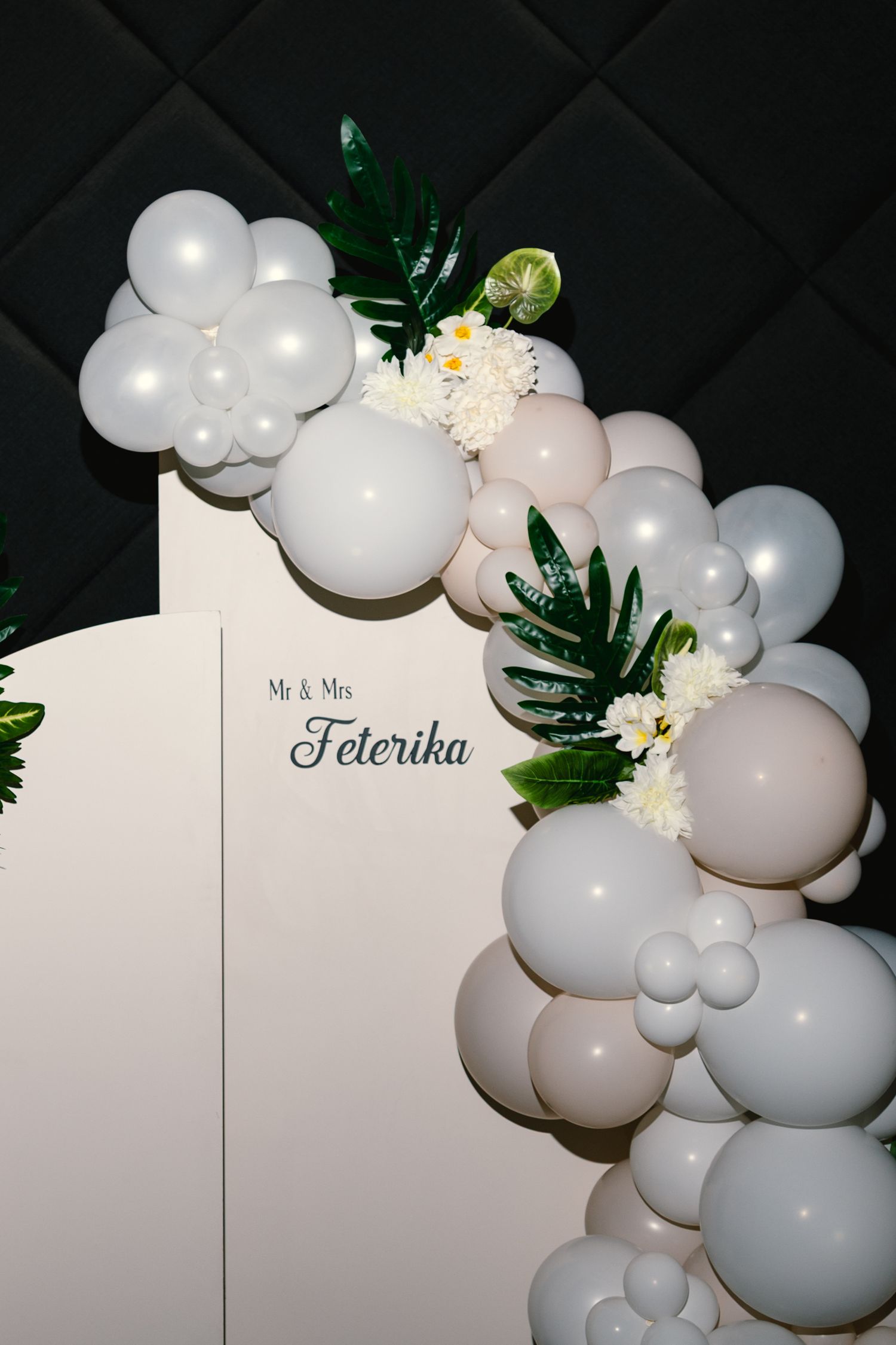 Elegant white balloon arch with tropical greenery and floral accents against a white backdrop for a wedding celebration.