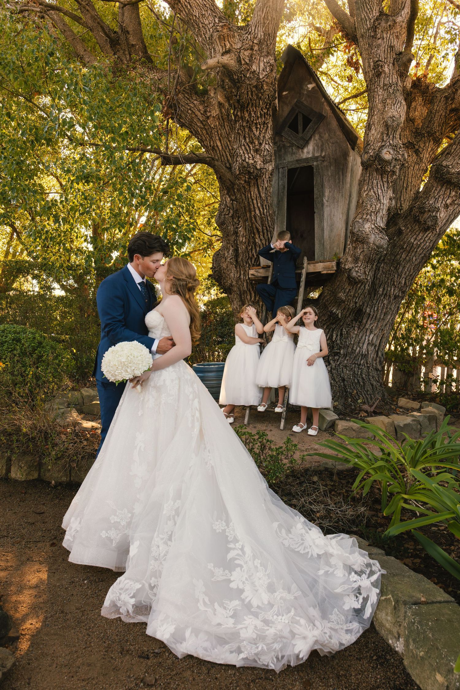 Bride and groom share a kiss in a garden with flower girls near a treehouse, bride in lace ball gown holding white bouquet.