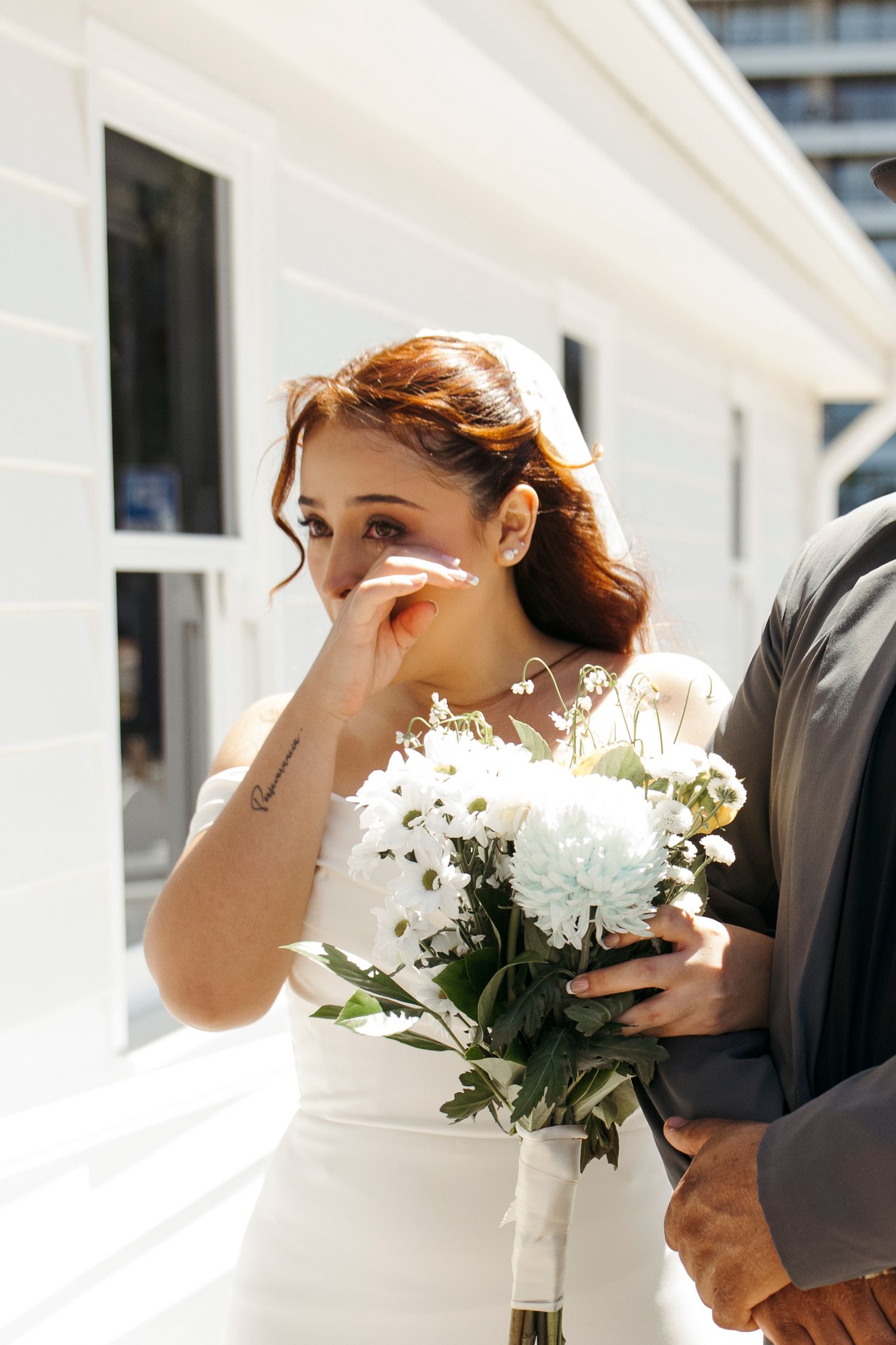 A bride in a white dress holds a bouquet of white flowers while wiping tears from her eyes during an emotional moment.