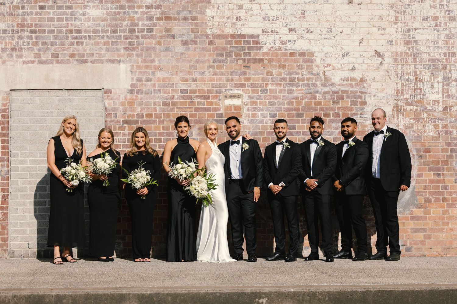 Wedding party in formal black attire poses against rustic brick wall for elegant group photo.