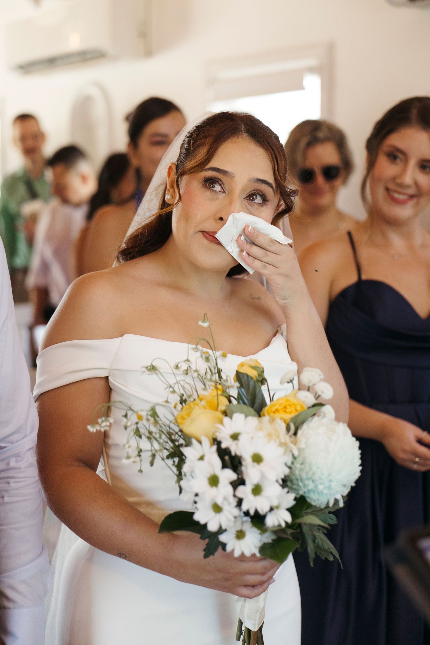 Bride in off-shoulder white dress wipes tears while holding yellow and white bouquet during emotional wedding moment.