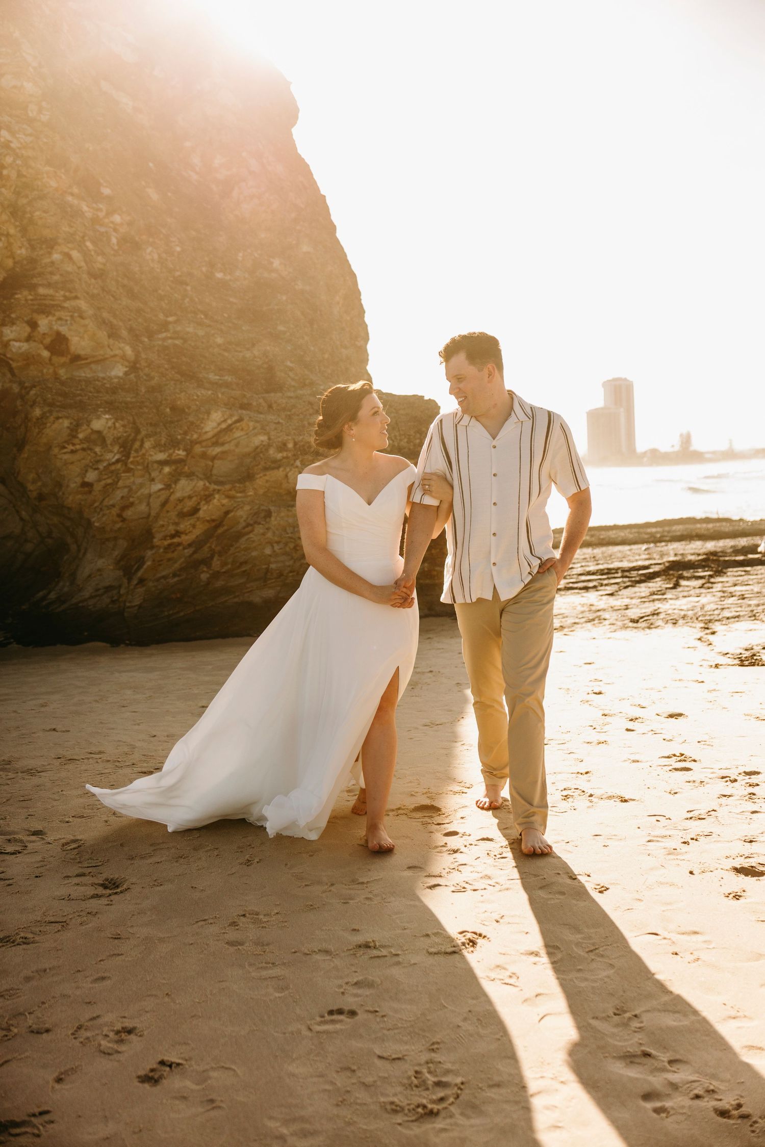A couple in wedding attire shares intimate moments on a sunlit beach with dramatic coastal rock formations.
