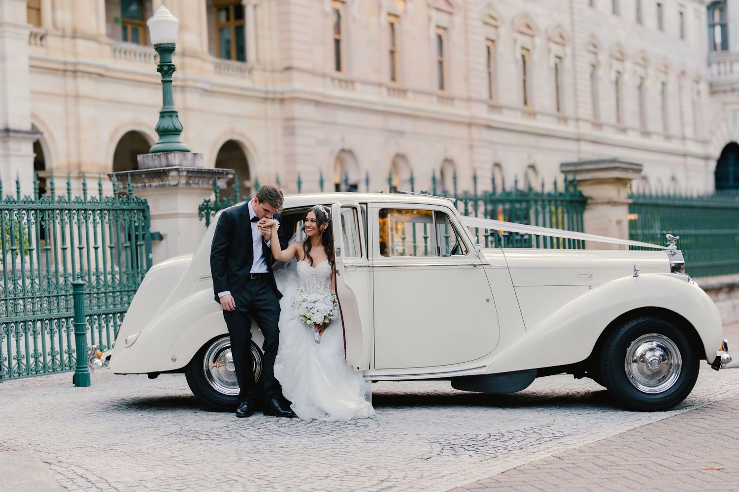 Bride and groom posing beside a vintage white Rolls-Royce with ribbon, outside a grand European building.