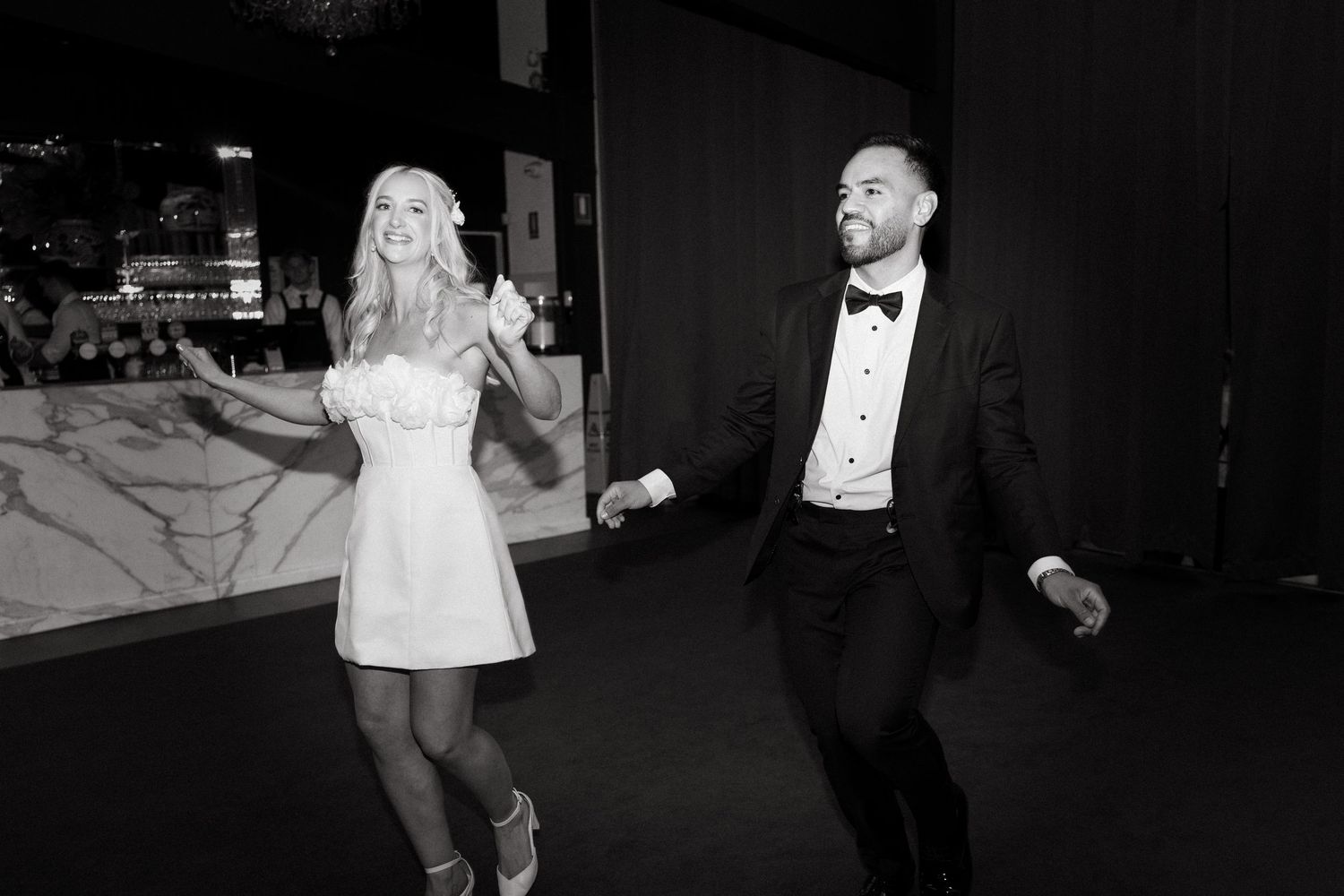 A couple in formal attire dances together on a dark street at night, captured in black and white photography.