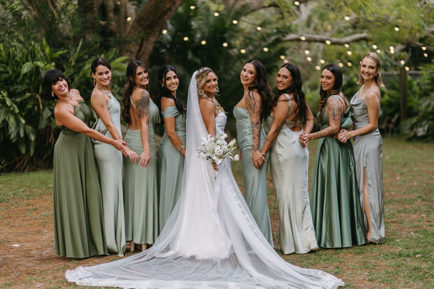 Wedding party wearing sage green and white dresses poses together in an outdoor garden setting.