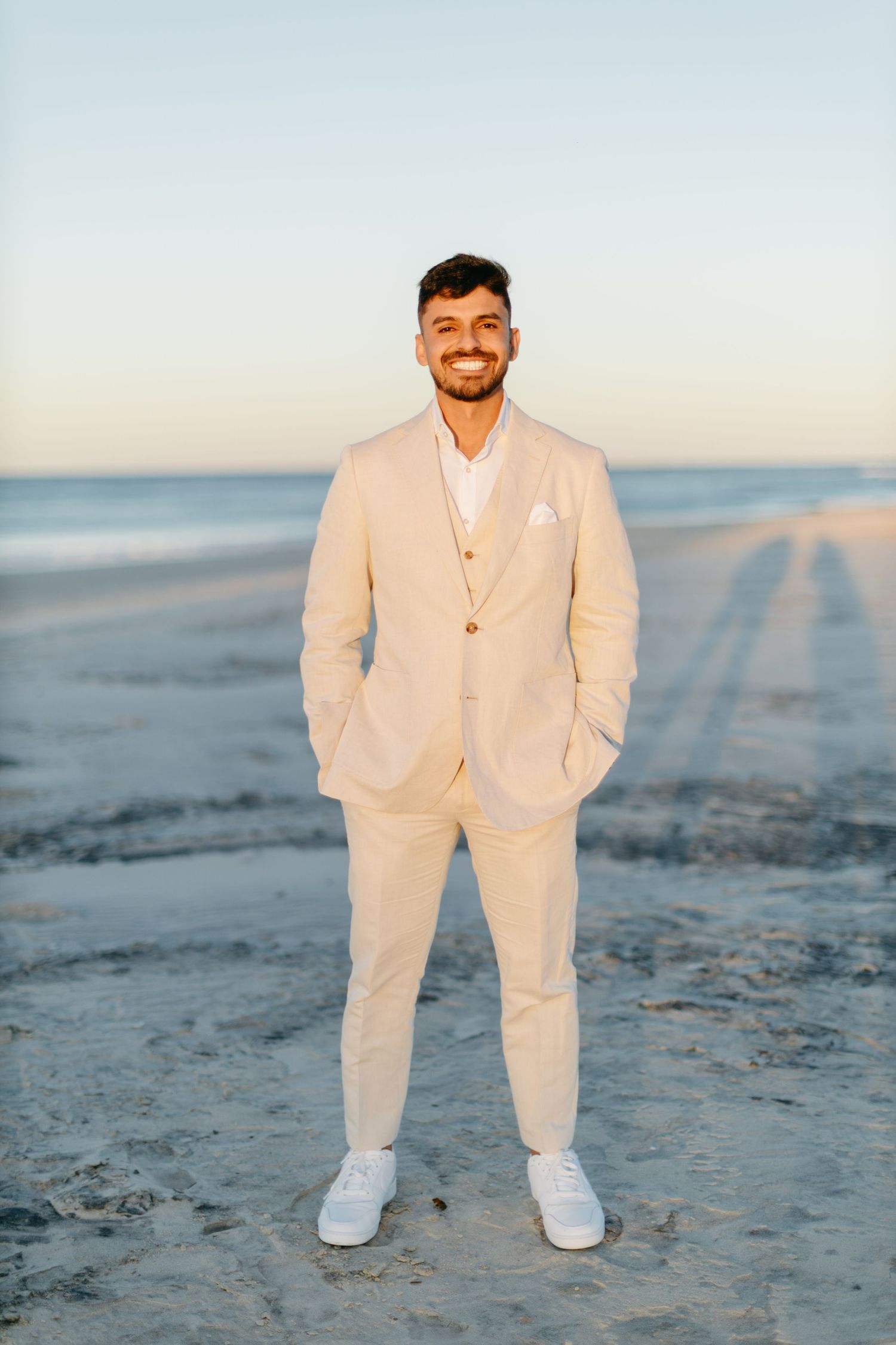 Groom in cream colored suit and white sneakers poses on sandy beach at sunset.