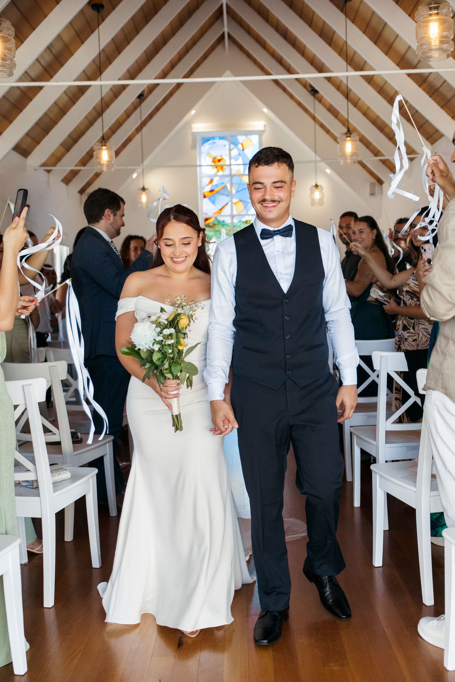 A wedding couple and guests laugh and dance together in a bright chapel with white chairs and vaulted wooden beams.