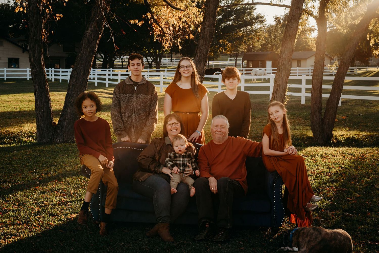 A multi-generational family portrait taken outdoors with white fencing and golden sunlight.