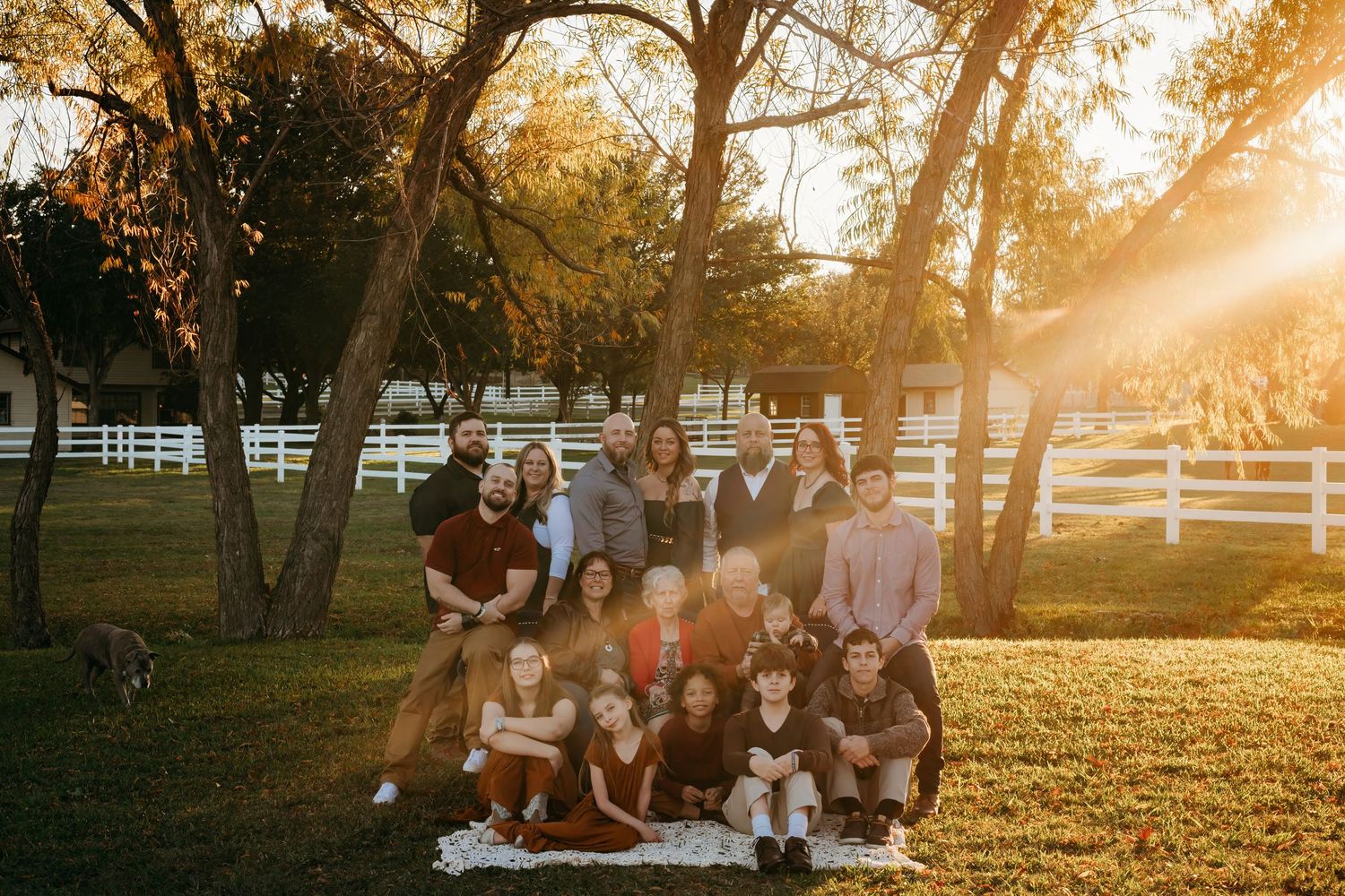 A large family gathering poses for a group photo at sunset with white fencing and trees.