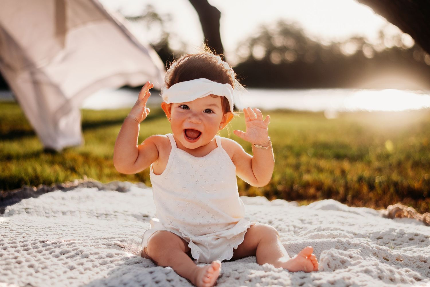 A happy baby in white clothing raises hands while sitting on a blanket outdoors.