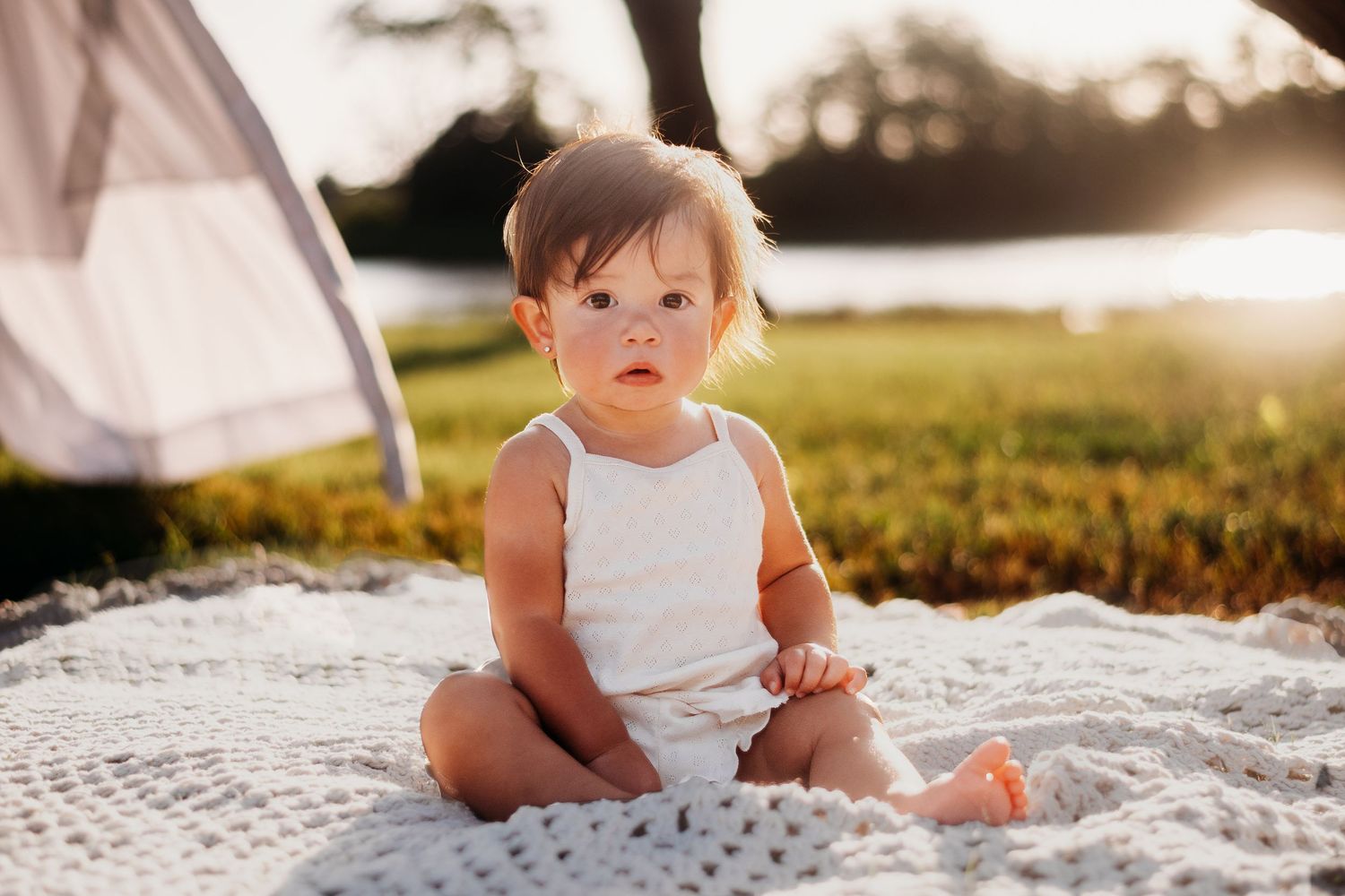 A baby in white clothing sits on a crocheted blanket during an outdoor photoshoot.