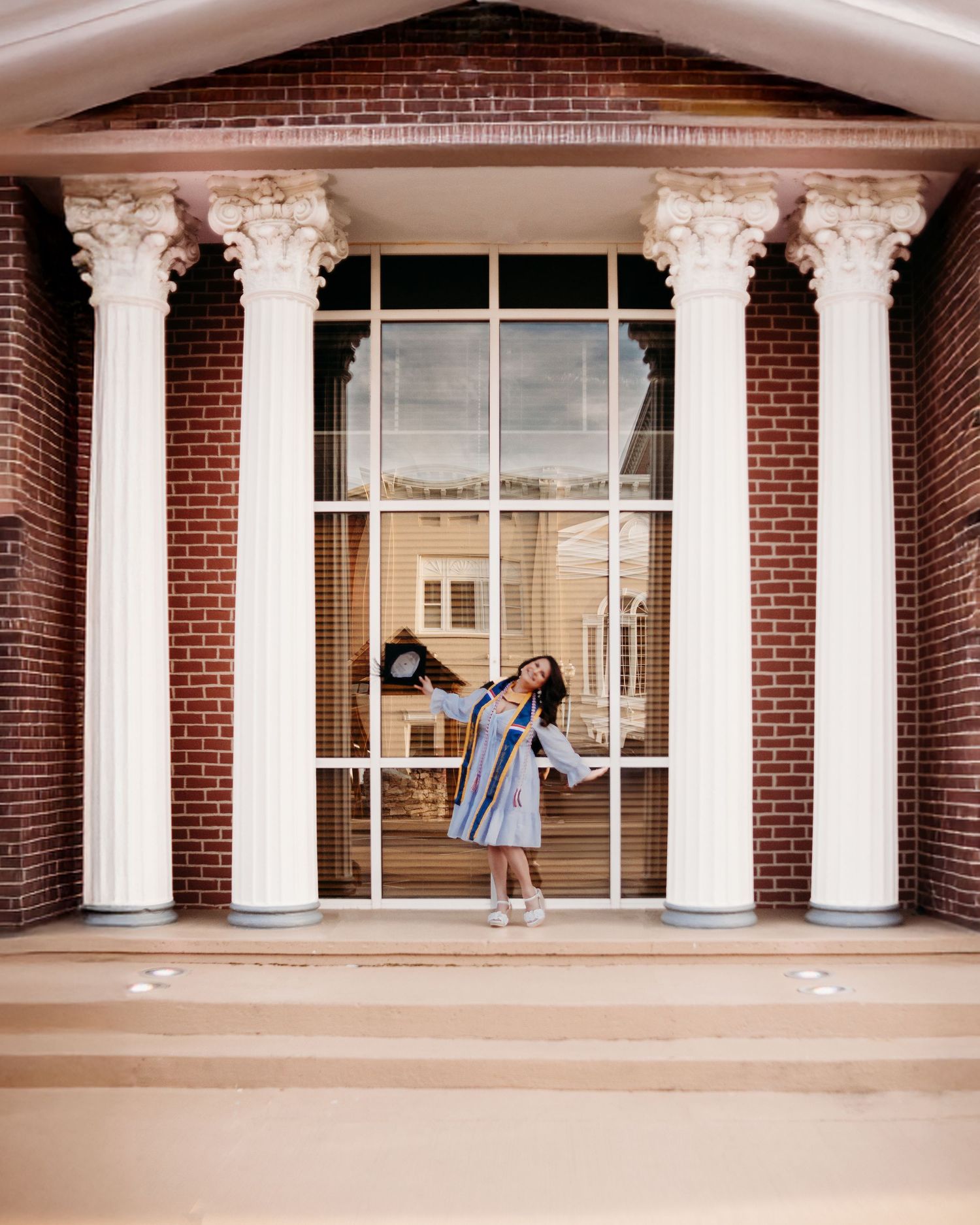 A graduate celebrates between white columns of a classic brick building entrance.