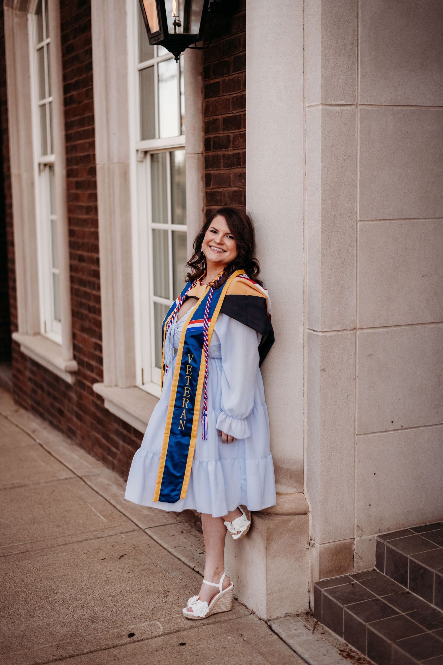 A graduate poses in academic regalia against a brick building.